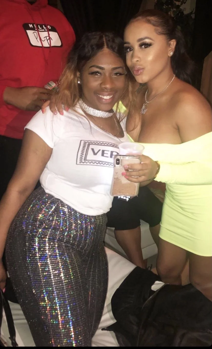 Two women at a social gathering, smiling and posing for a photo. One woman is wearing a white graphic T-shirt and shiny, patterned pants, holding a drink and a phone. The other woman is dressed in a bright yellow off-shoulder dress, holding a drink, 