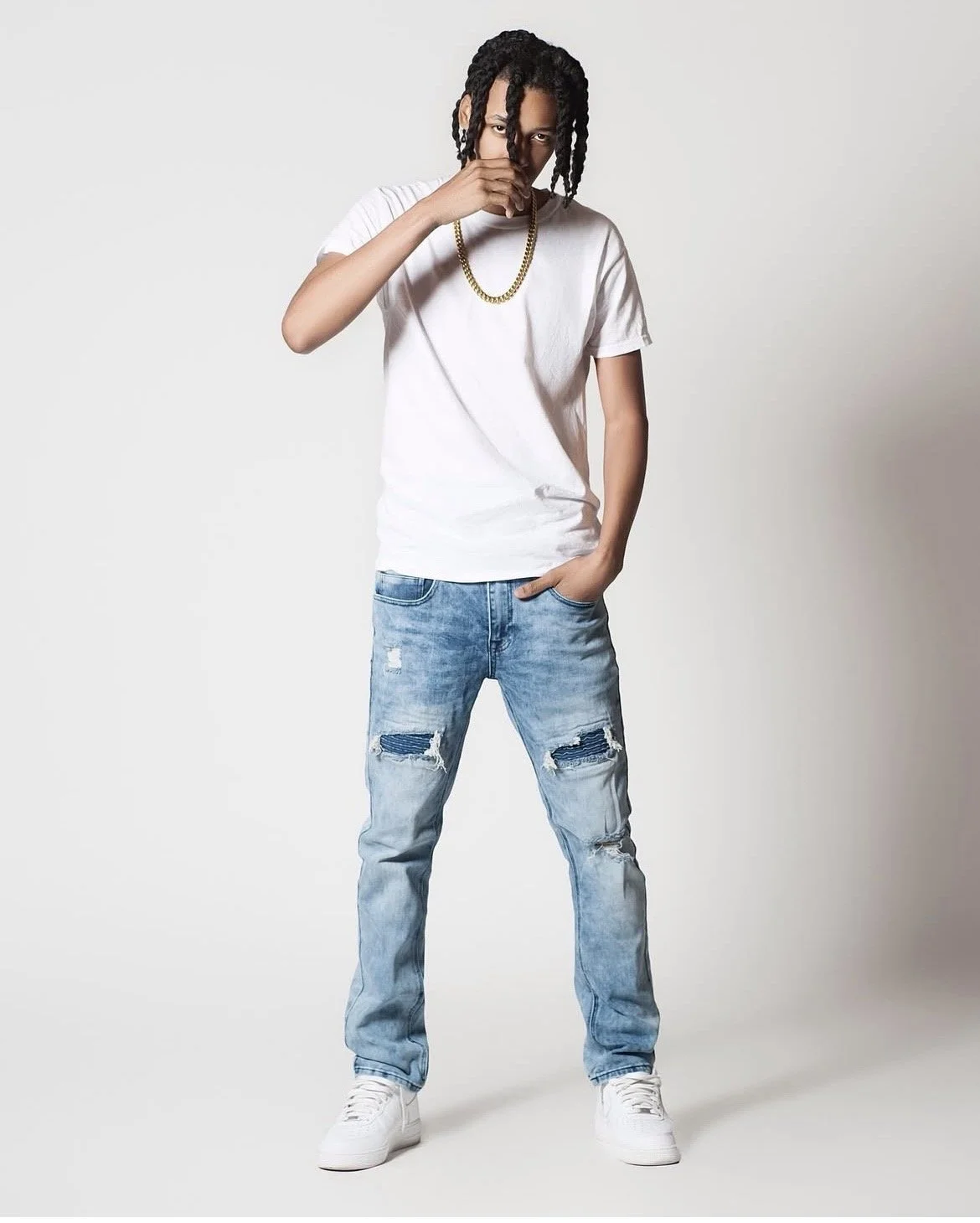 A young man with braided hair, wearing a white t-shirt, ripped jeans, white sneakers, and a gold chain, standing against a plain white background, with one hand in his pocket and the other hand near his face.