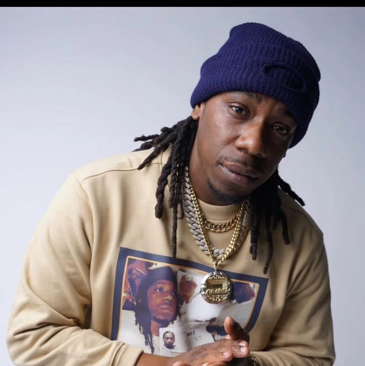 A man with dreadlocks wearing a navy beanie, gold chains, and a beige sweatshirt with multiple pictures printed on it, posing against a light gray background.