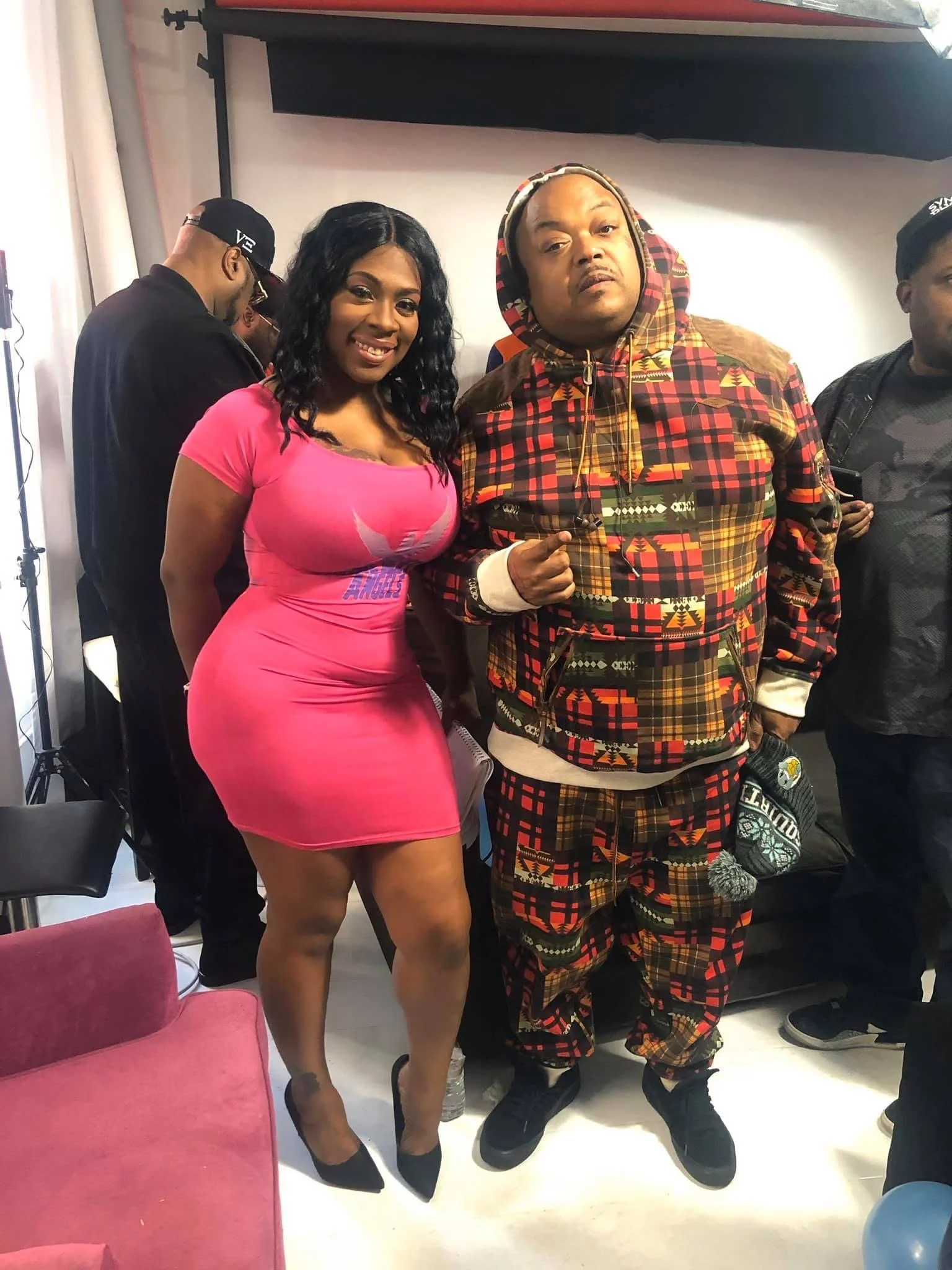 A woman in a tight pink dress standing next to a man in a colorful, patterned hoodie and matching pants at an indoor event.