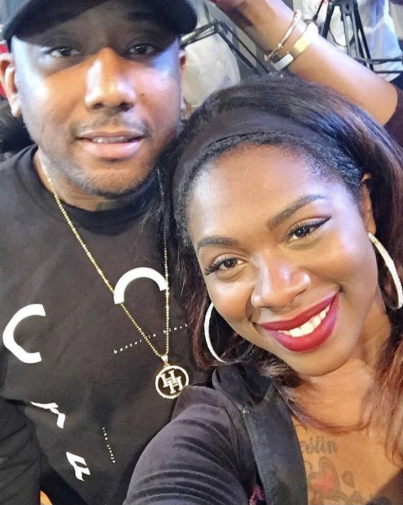 A man and woman taking a selfie together, smiling. The man is wearing a black cap, a black t-shirt, and a gold chain with a pendant. The woman has long dark hair, large hoop earrings, red lipstick, and a tattoo visible on her chest.