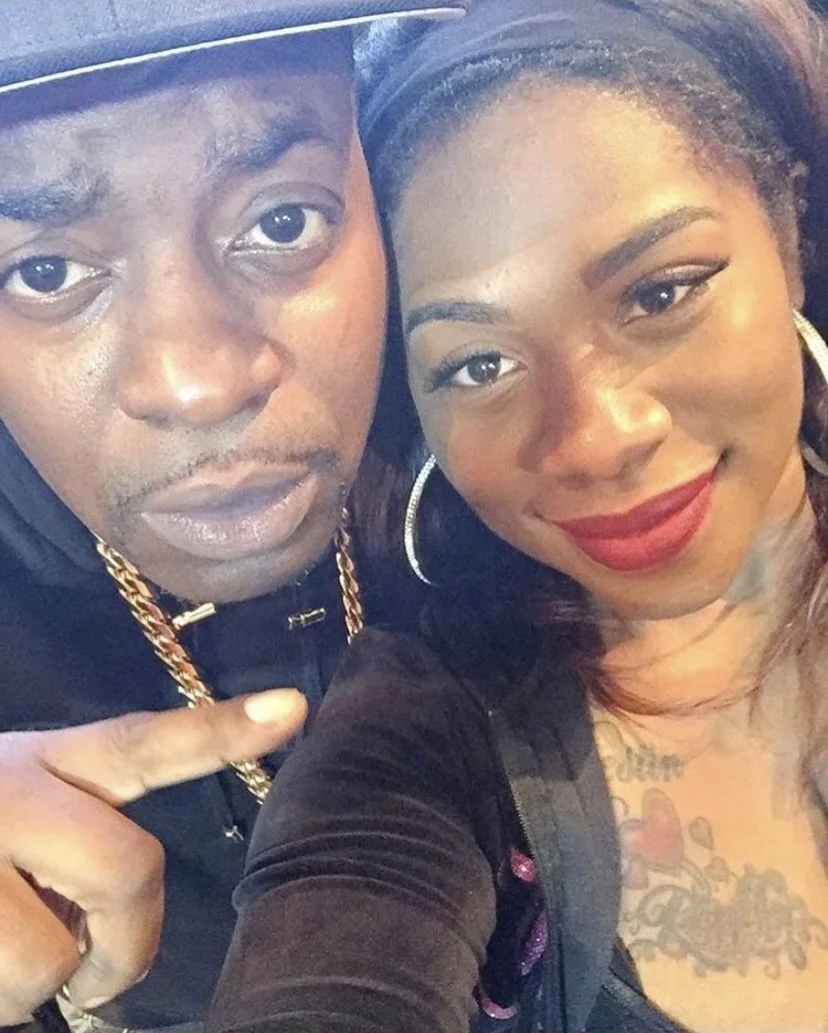 A close-up of a man wearing a baseball cap and a woman with red lipstick and hoop earrings, both looking at the camera.