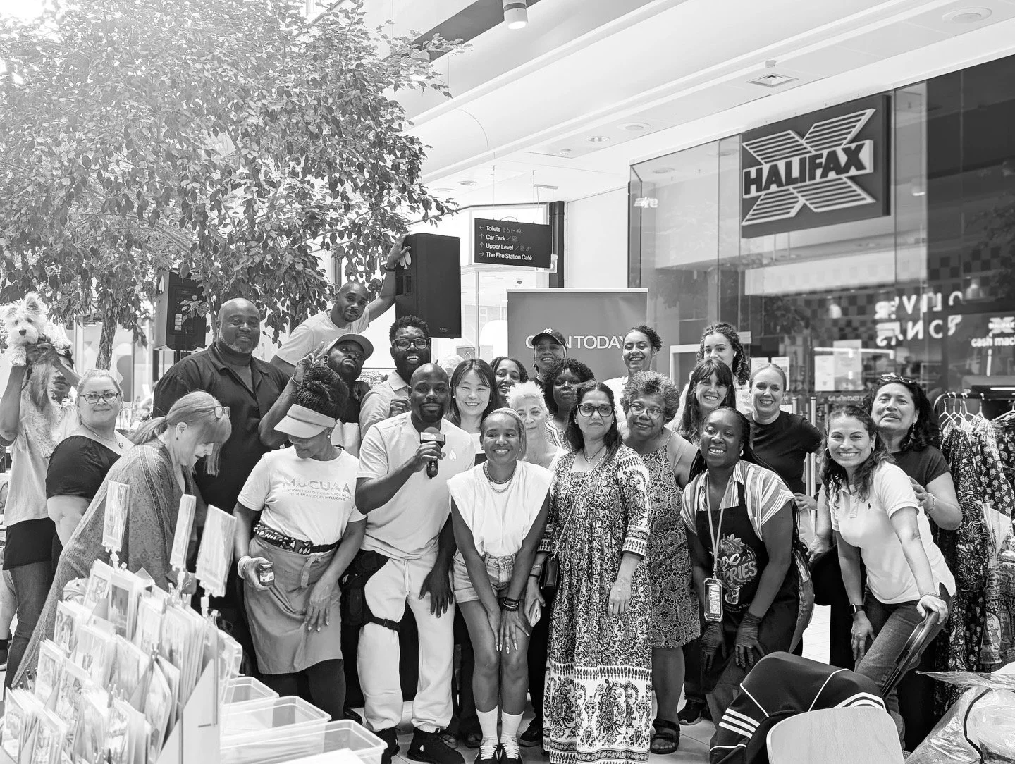 Group of diverse people smiling and posing together inside a shopping mall, with a store sign 'HALIFAX' and display racks in the background.