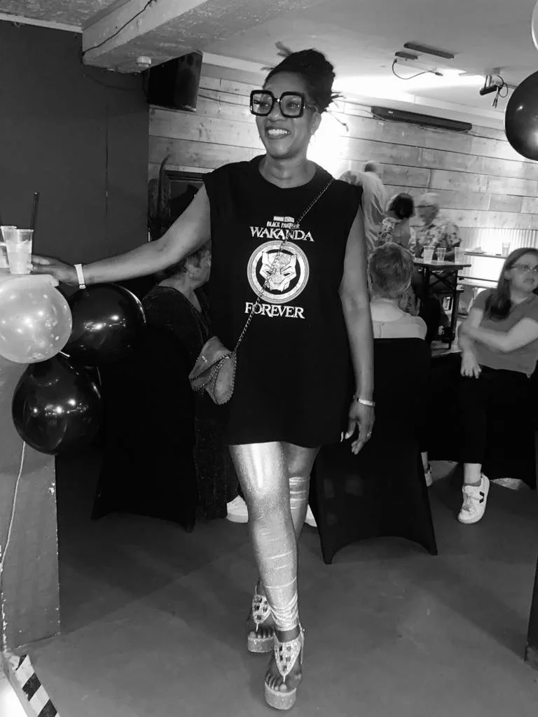 A woman smiling at a party, wearing large glasses, a black 'Black Panther Wakanda Forever' T-shirt, shiny leggings, and platform shoes, standing near balloons and other guests in a social setting.