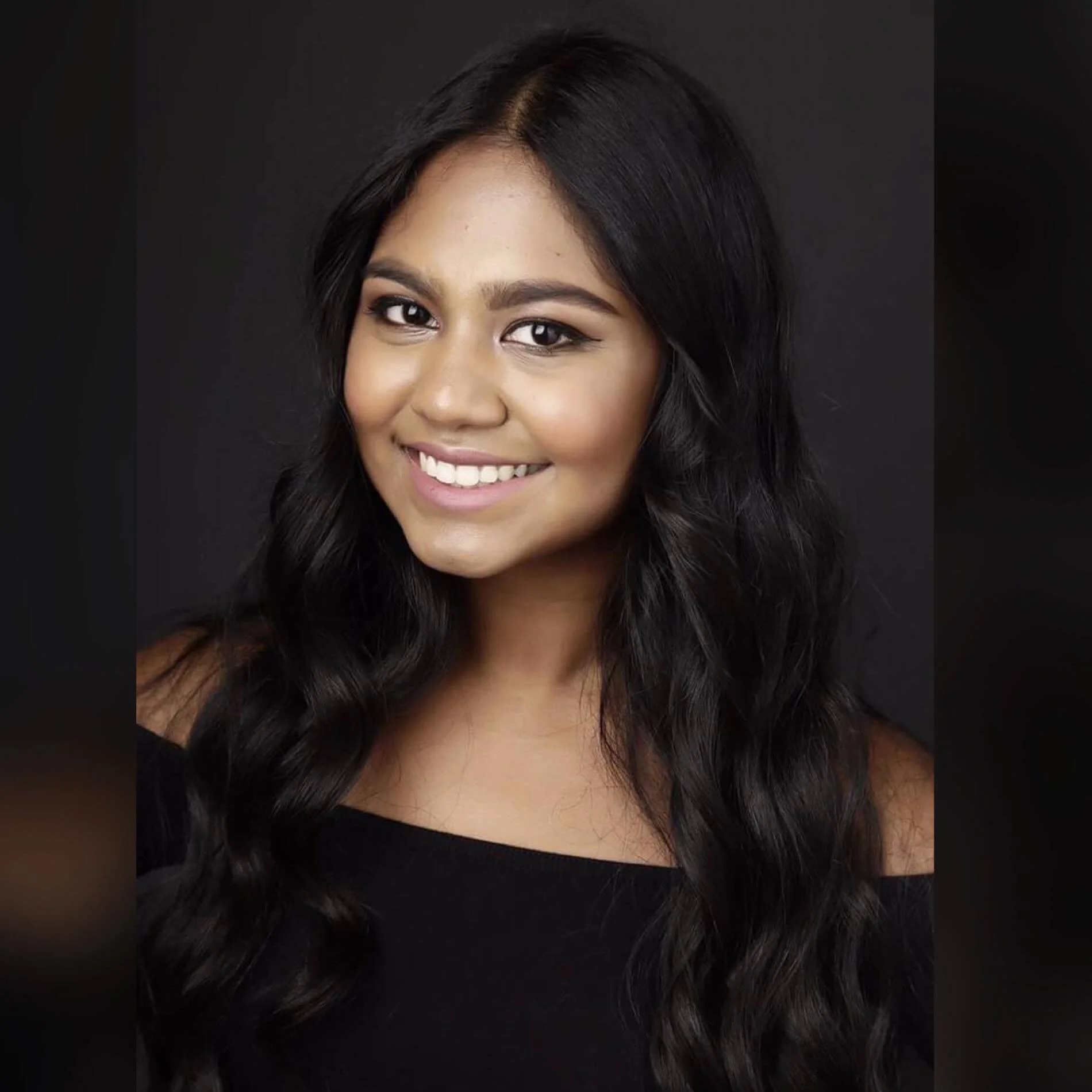 SOPHIA ESTELLE. A woman with long black hair styled in loose waves, smiling, wearing a black top, against a dark background.