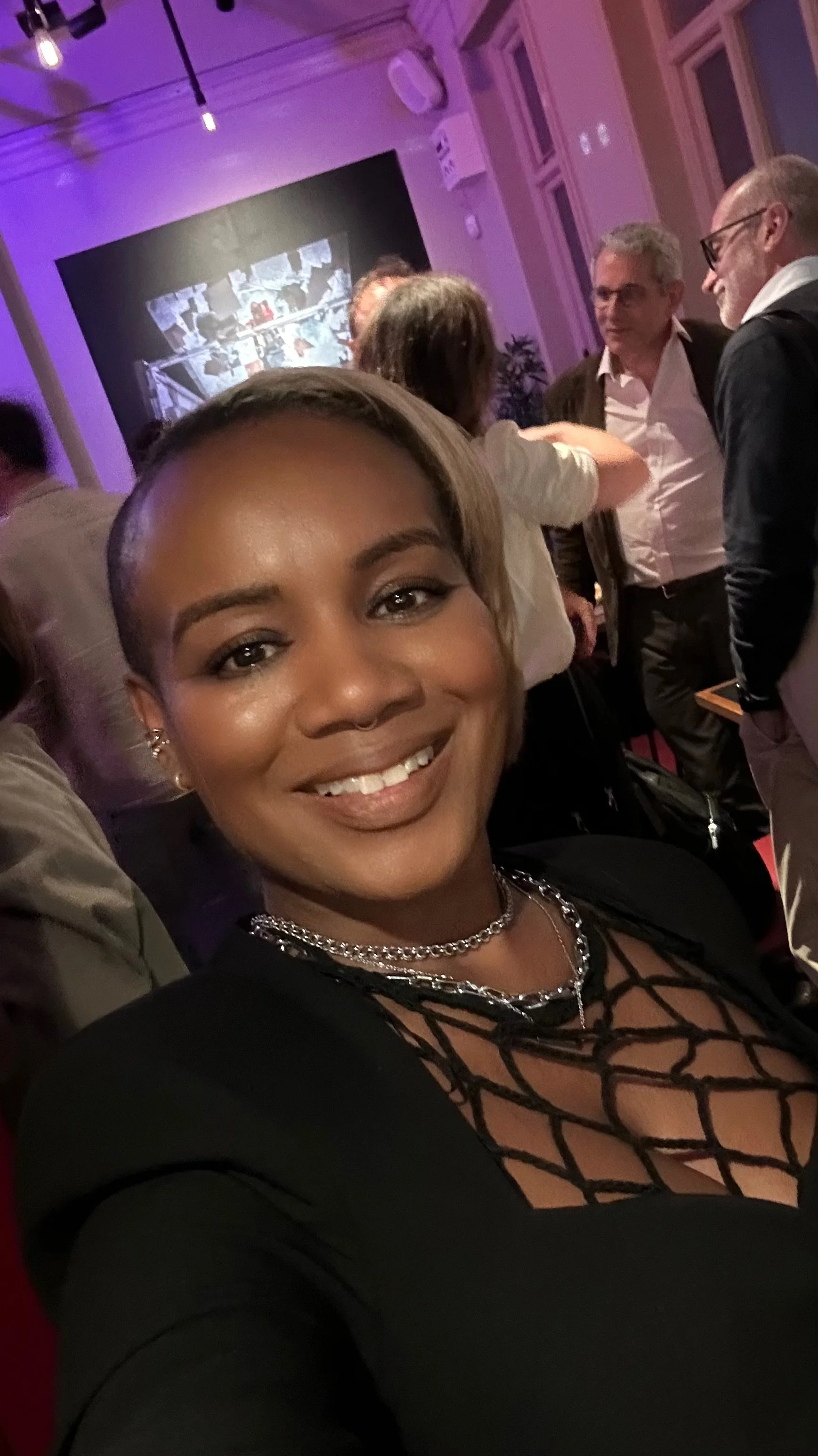 A ISHA NGABILE with a shaved head smiling at the camera, wearing layered necklaces and a black dress with a lace pattern on the chest, at a crowded indoor event with purple lighting.