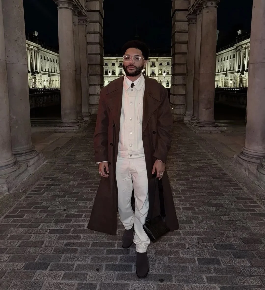 Man standing outdoors at night in front of historical building with stone columns, wearing glasses, a tan trench coat, a light-colored jacket and pants, holding a bag.