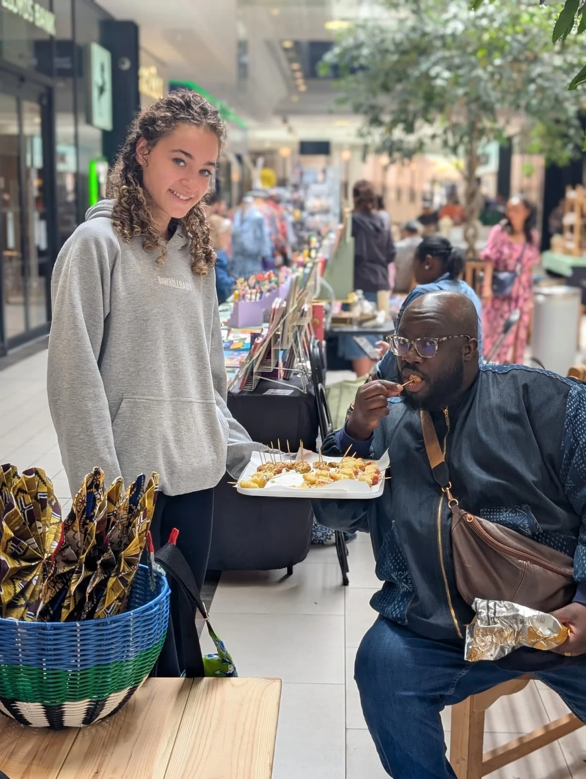 A young woman with curly hair, wearing a gray hoodie, smiling while standing at a food stall. A man with glasses, eating food from a paper tray with skewers, seated nearby. In the foreground, a basket filled with black and yellow patterned napkins or cloths. The background shows a busy indoor market with several people shopping and walking around.