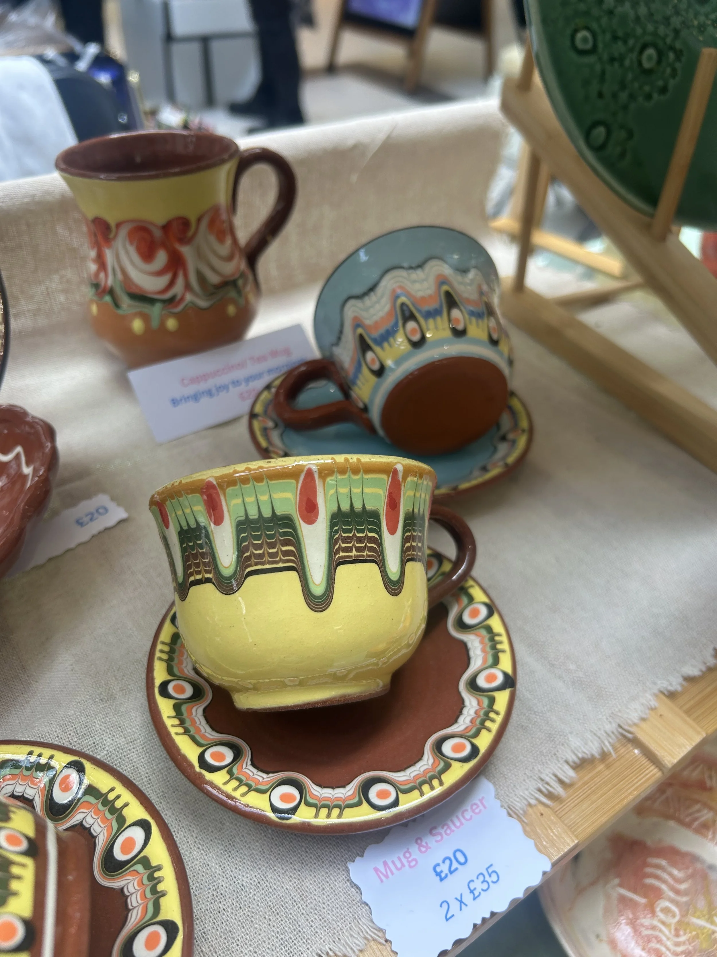 Colorful ceramic mug and saucer set with an abstract pattern, displayed on a table at a market or craft fair.