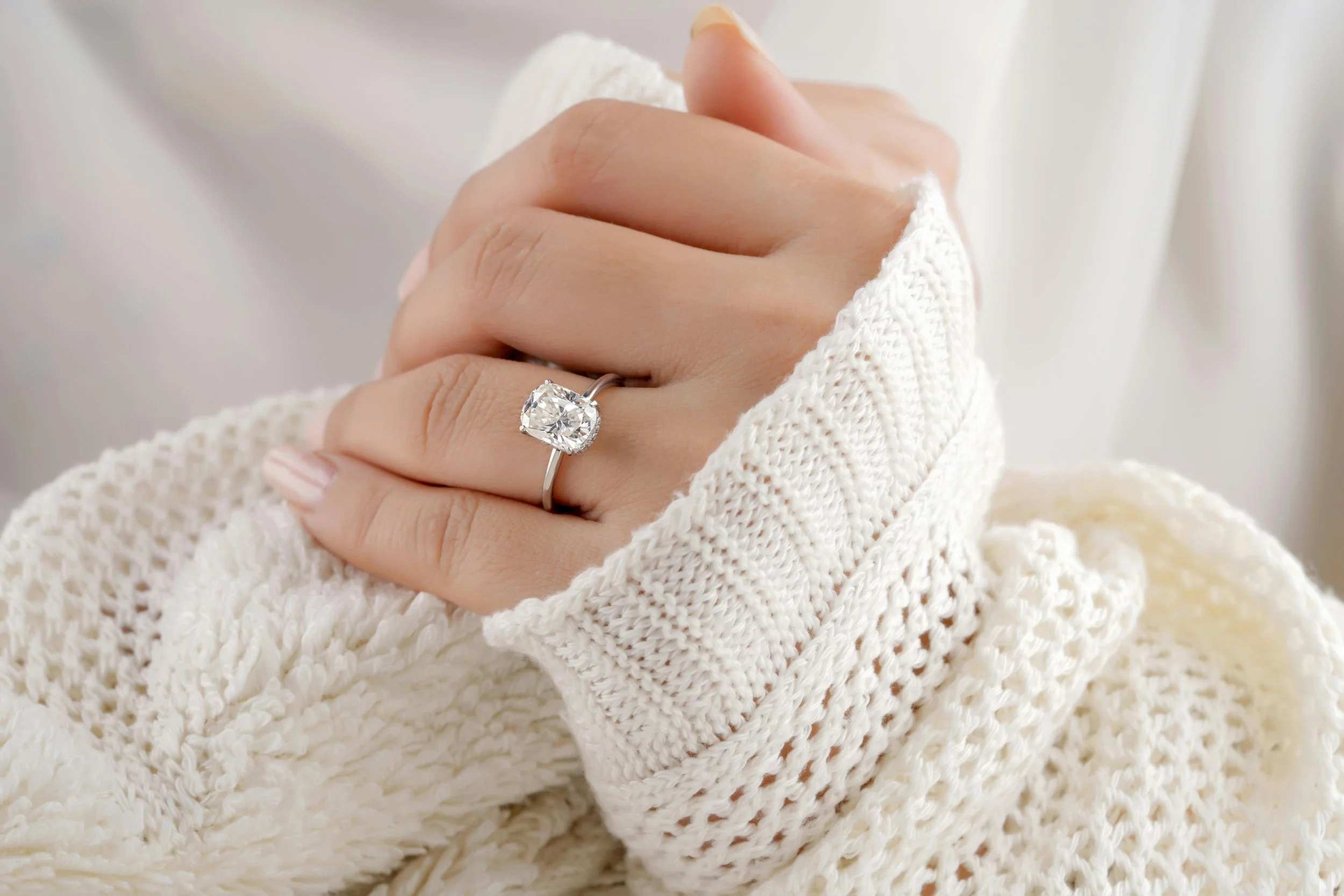 Close-up of a woman's hand wearing a diamond engagement ring on her ring finger, with her hand resting on a knitted white sleeve.
