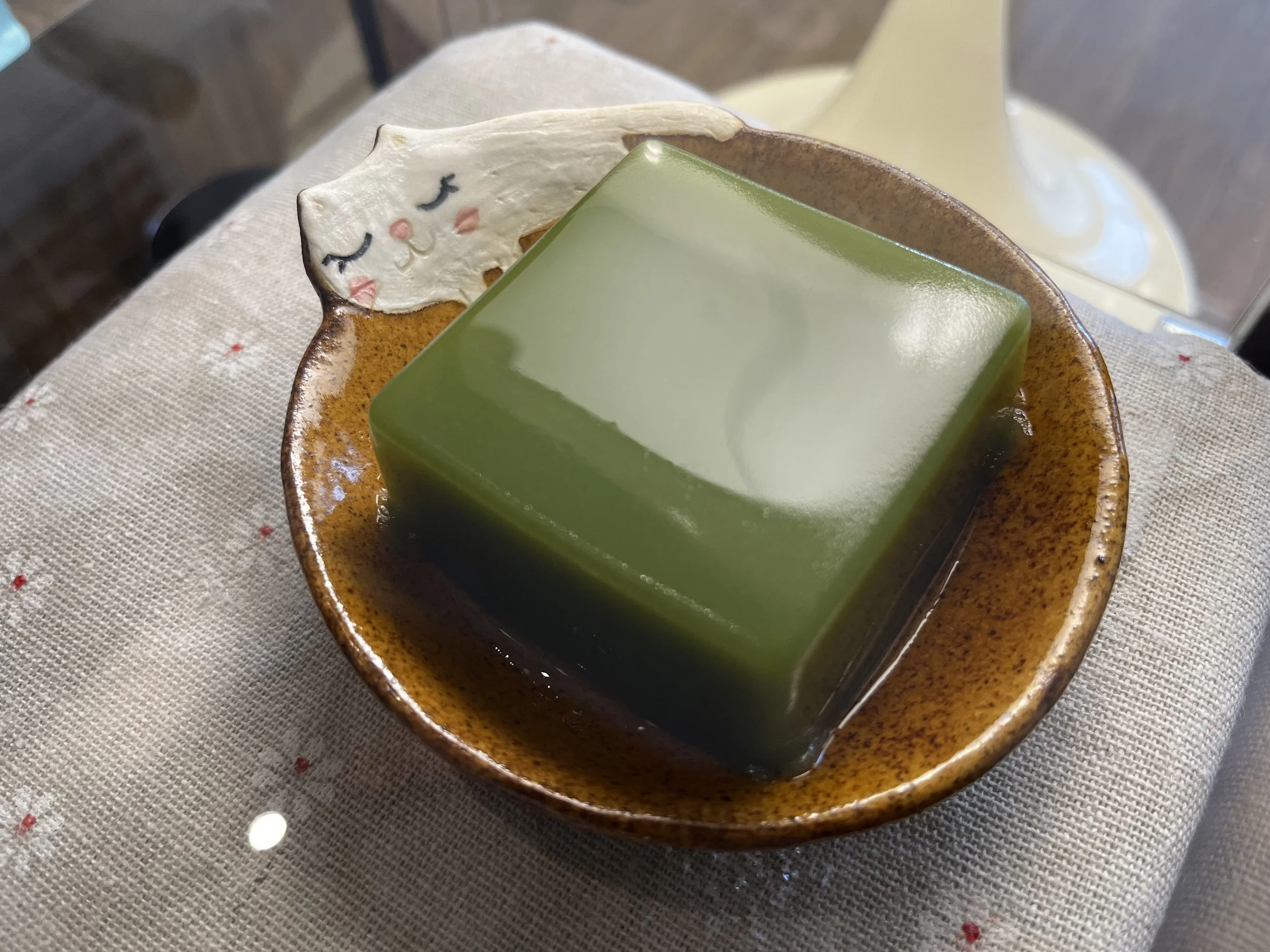 A green cube-shaped jelly dessert on a brown ceramic dish with a small white animal-shaped decoration attached to the edge of the dish.
