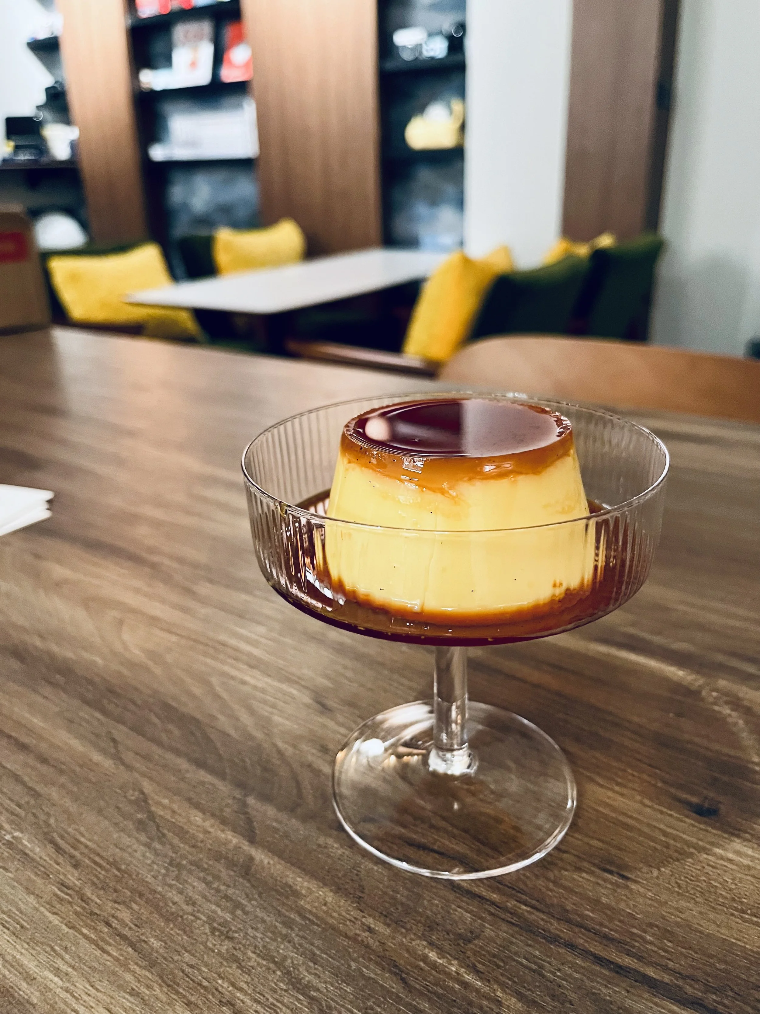 Caramel flan dessert in a glass cup on a wooden table with a blurred background of chairs and shelves.