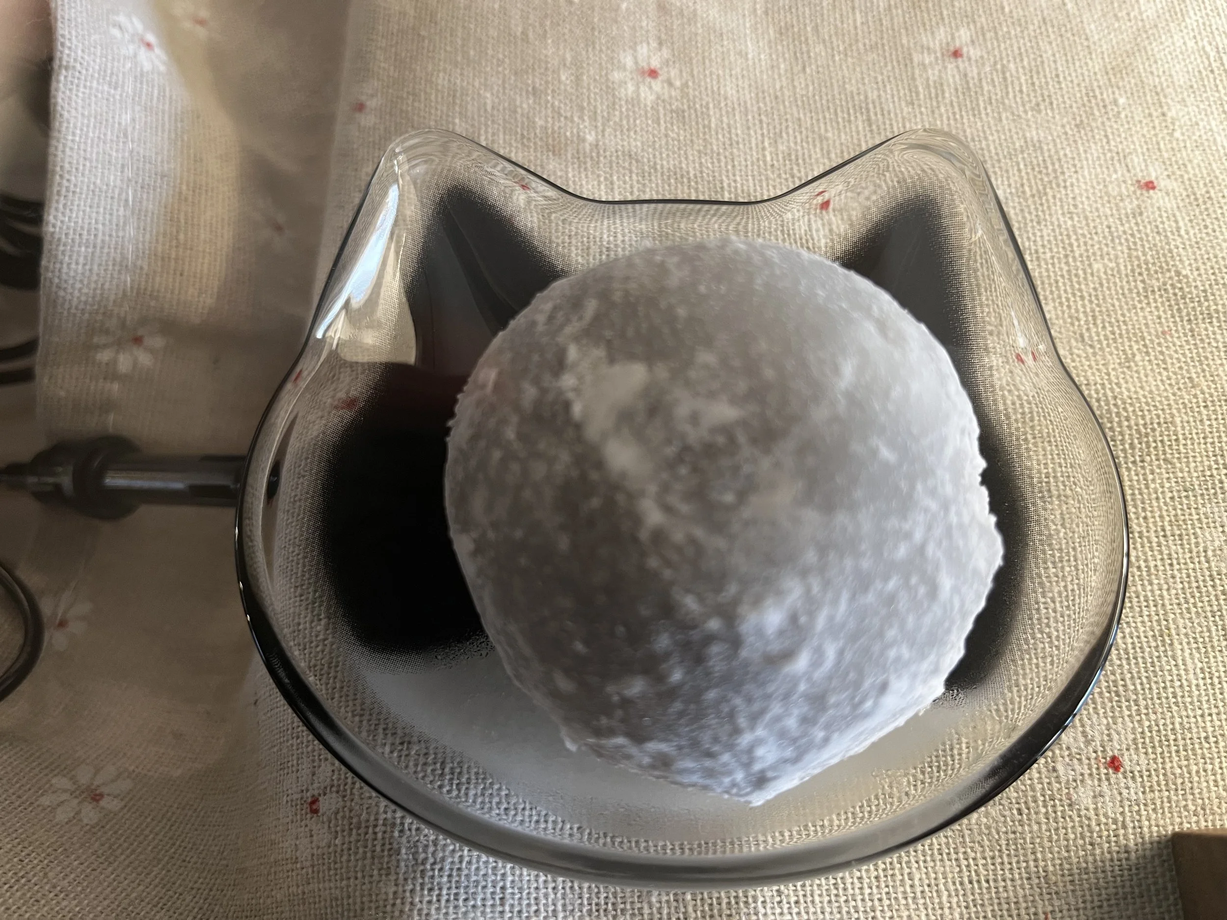 A glass bowl contains a white, powdery snowball on a table with a beige tablecloth.