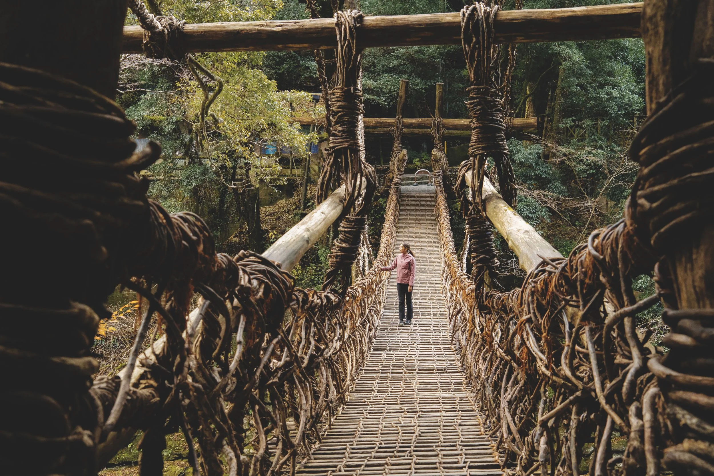 A woman in a pink jacket walking across a wooden suspension bridge made of logs and ropes, surrounded by green trees in a forest.