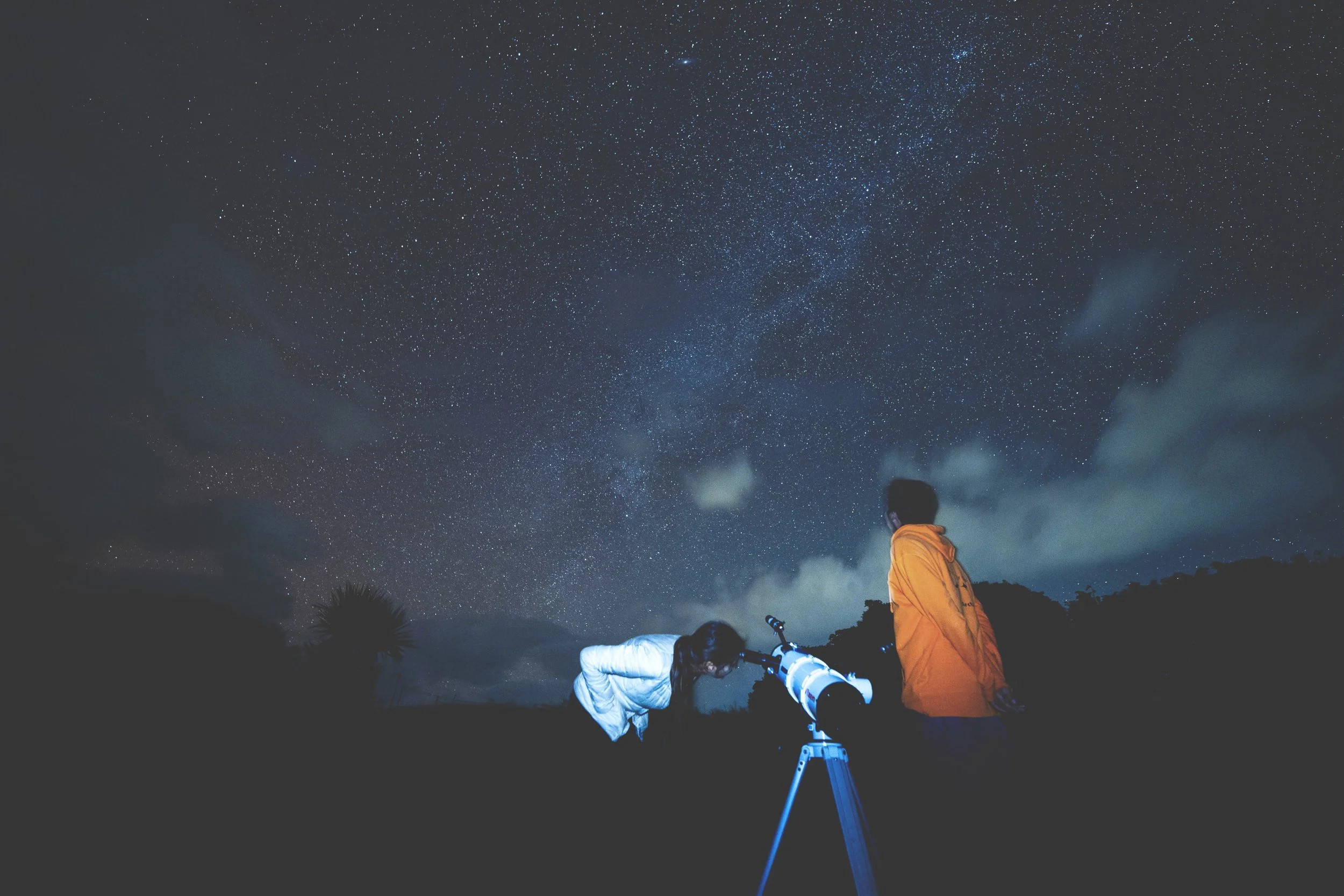 Two children, one in a white jacket and the other in an orange jacket, observing the night sky with a telescope outdoors, under a star-filled sky with clouds.