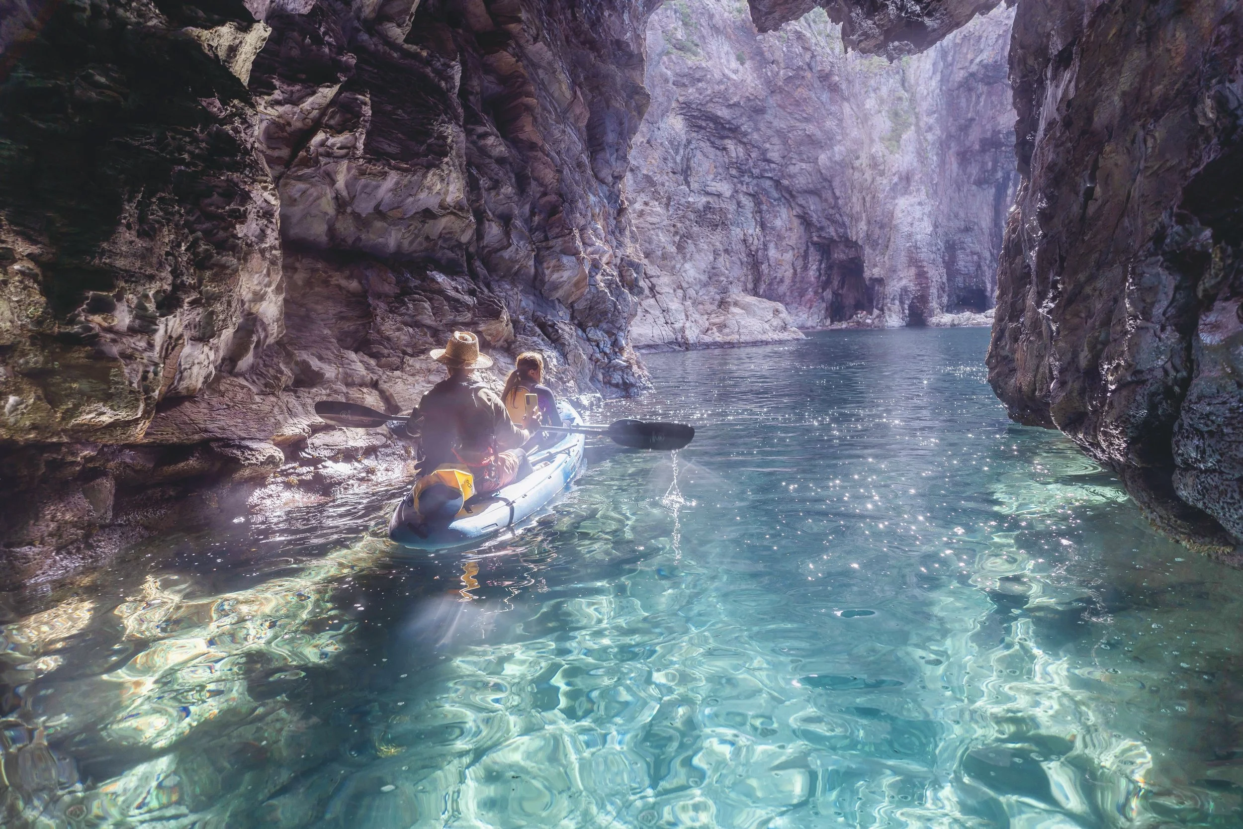 Two people kayaking in a narrow canyon with towering rocks and clear blue water.