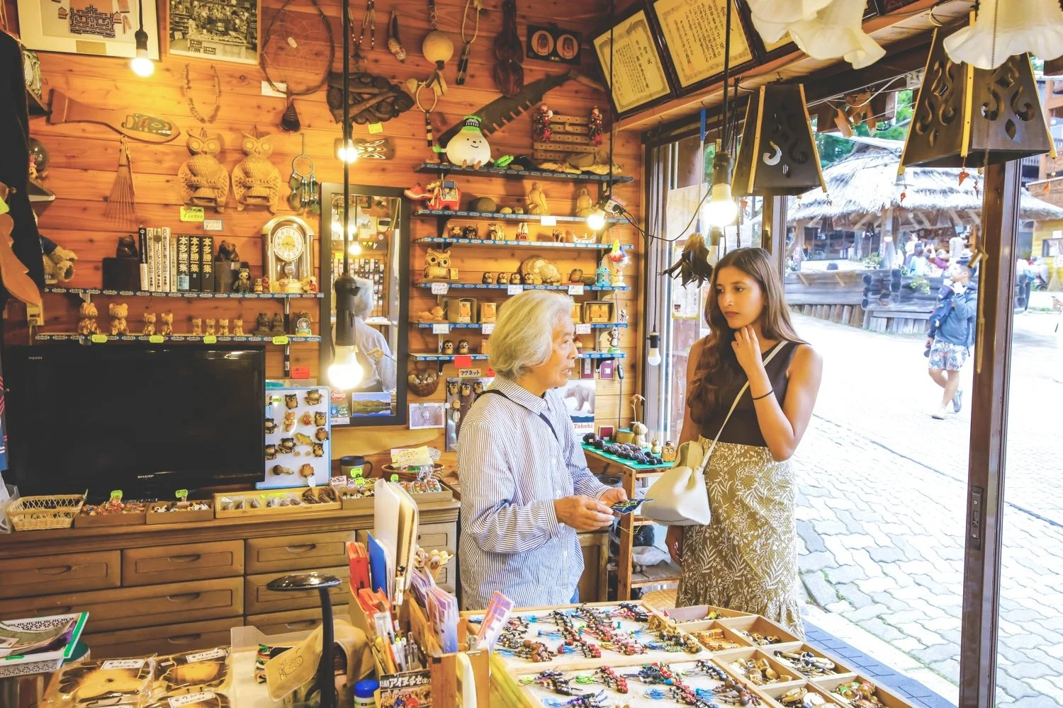 Inside a gift shop with wooden walls, two women are engaged in conversation; an older woman with gray hair and a younger woman with long brown hair. The shop displays various souvenirs, figurines, and jewelry, with shelves and counters full of small 
