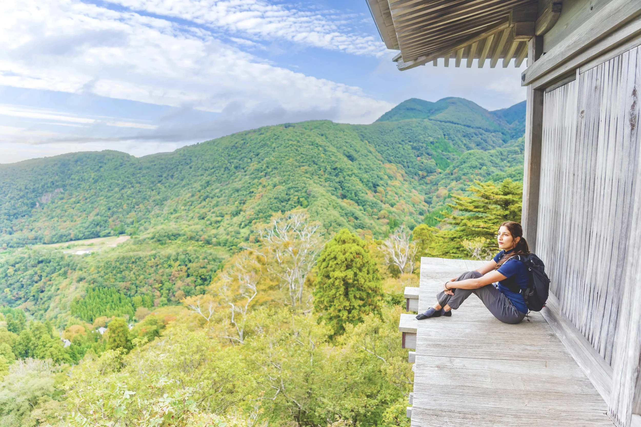 Young woman sitting on a wooden balcony overlooking lush green mountains and trees, wearing a backpack, blue shirt, and gray pants.