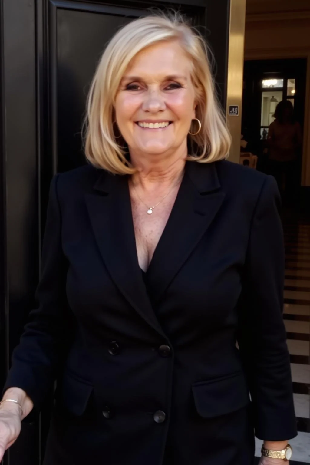 Smiling middle-aged woman with blonde hair, wearing a black blazer, gold jewelry, standing in front of a dark door and warmly lit interior.