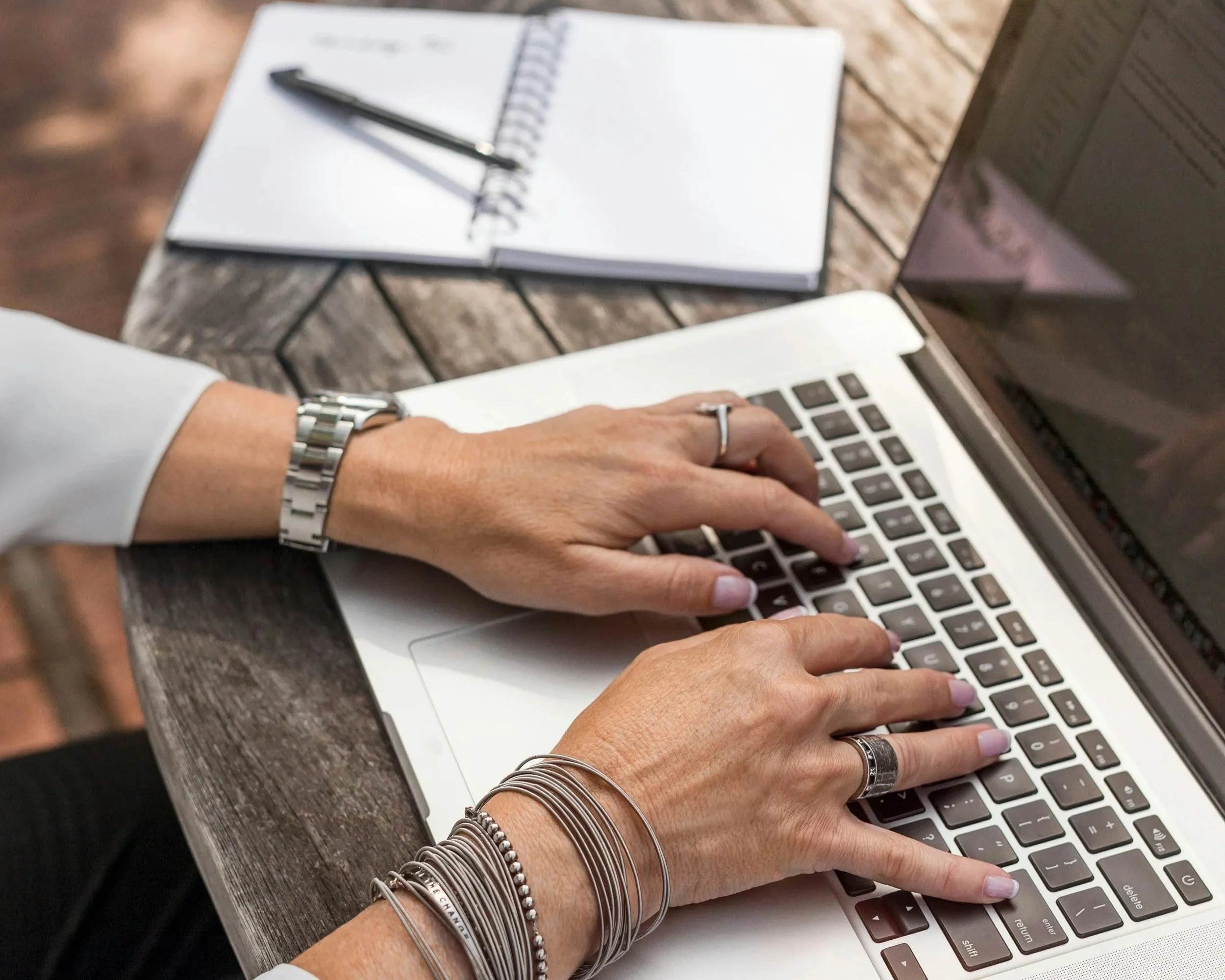 Woman's hands typing on a laptop keyboard on a wooden table, with a closed notebook and pen nearby.