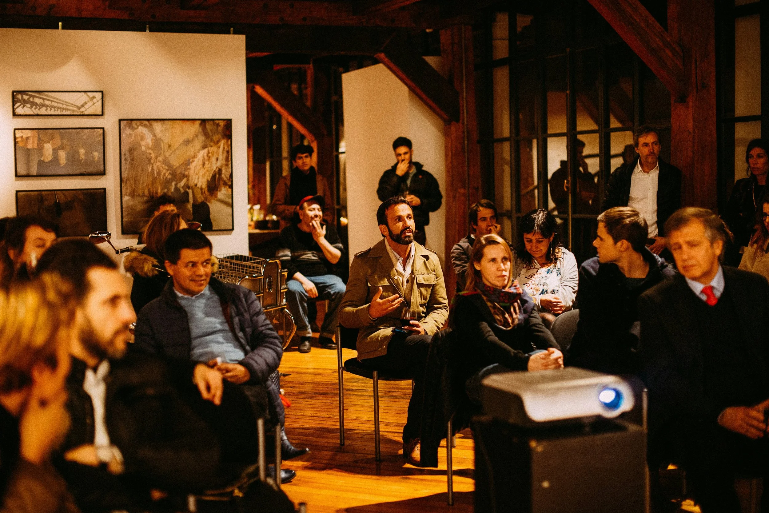 A group of people seated and standing in a warmly lit, rustic indoor space, possibly attending a presentation or talk, with art on the walls and wooden beams visible.