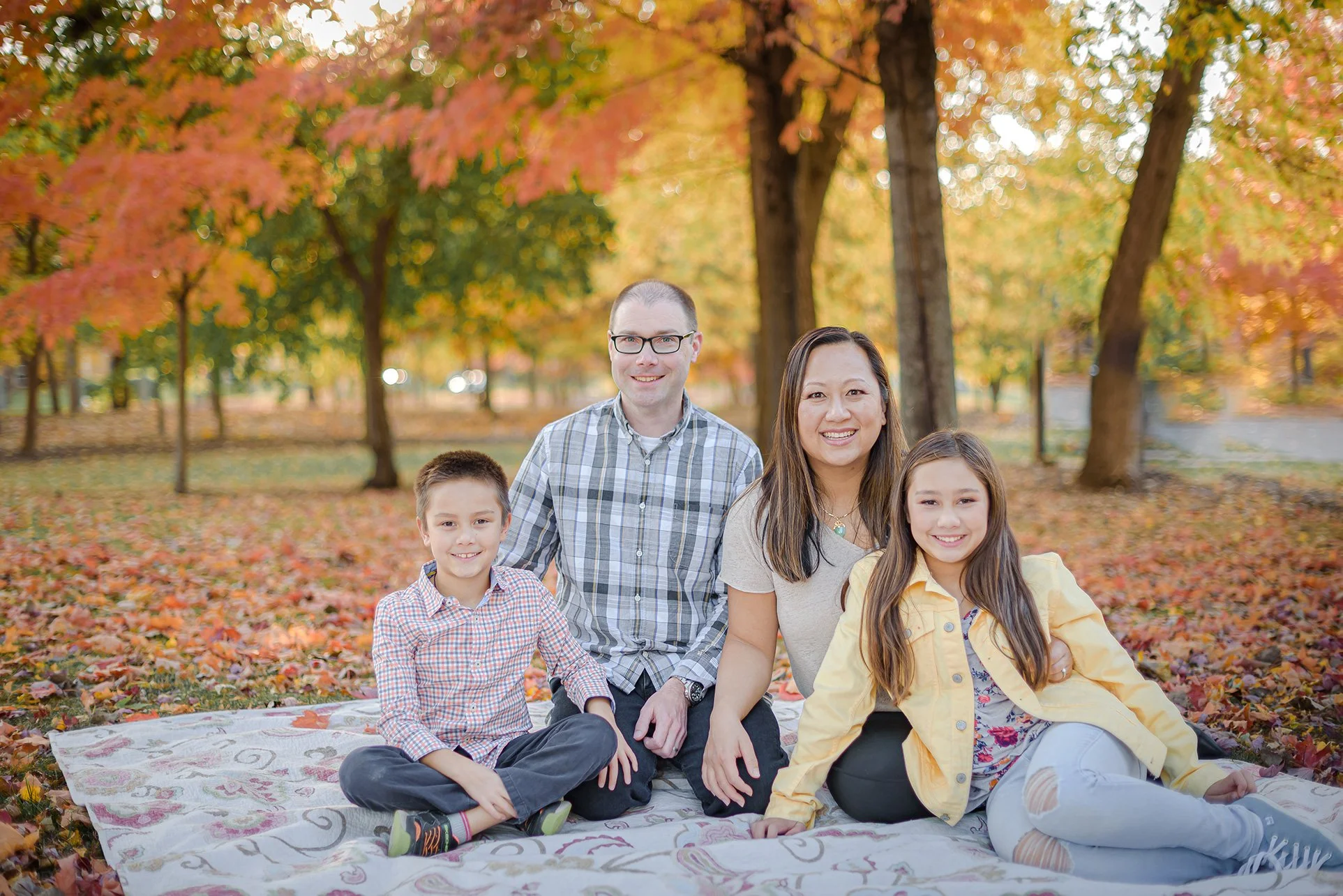 Henne family of four sitting on a blanket in Fraze Park, Kettering Ohio during fall, surrounded by colorful autumn trees and fallen leaves.