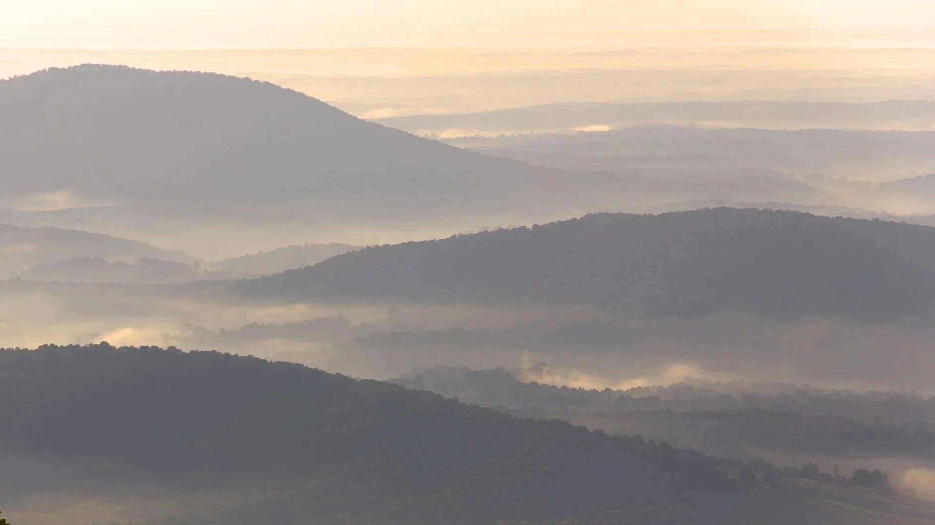 A landscape of multiple mountain ranges with layers of mist and fog, bathed in soft morning or evening light, creating a peaceful and ethereal scene.