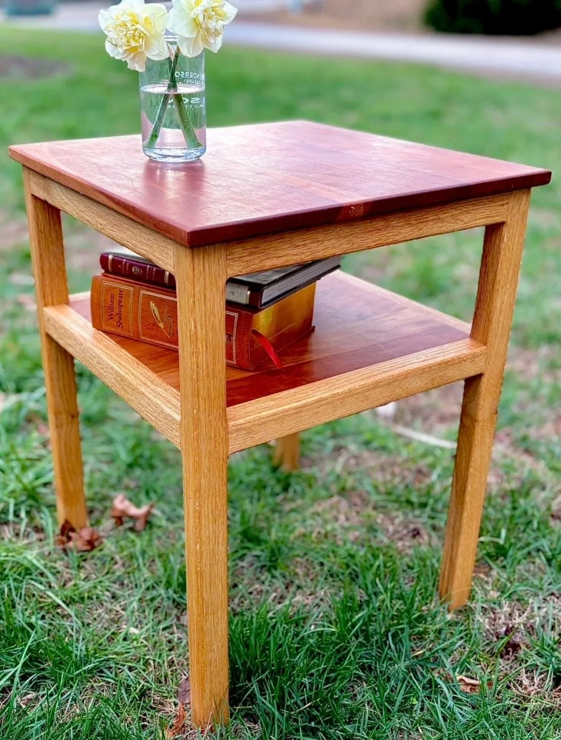 A wooden side table with a glass vase of white flowers on top and two books on the lower shelf, set outdoors on grass with a blurred background.