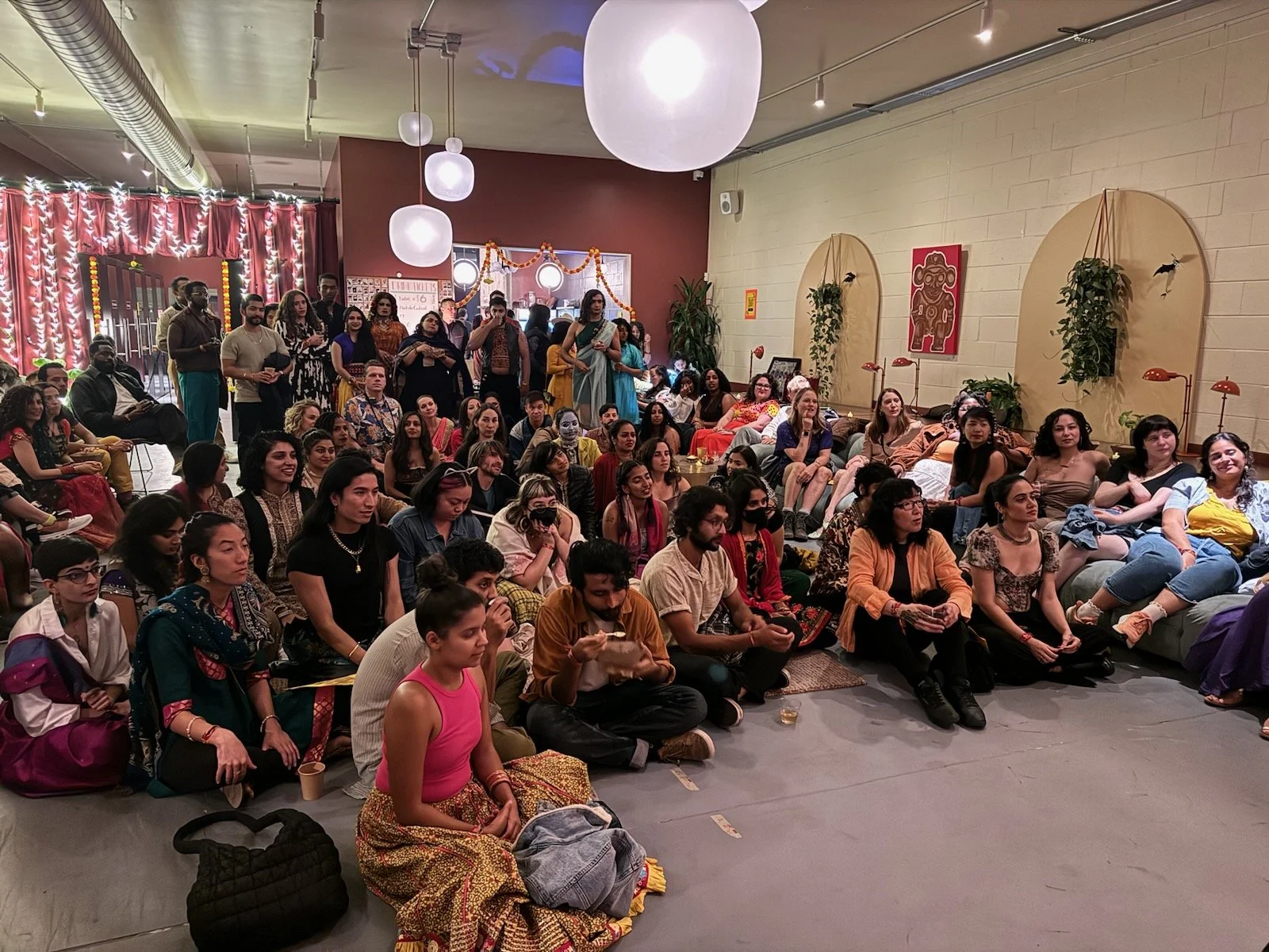 A large group of people seated and standing in a decorated indoor space, possibly attending a cultural event or gathering.