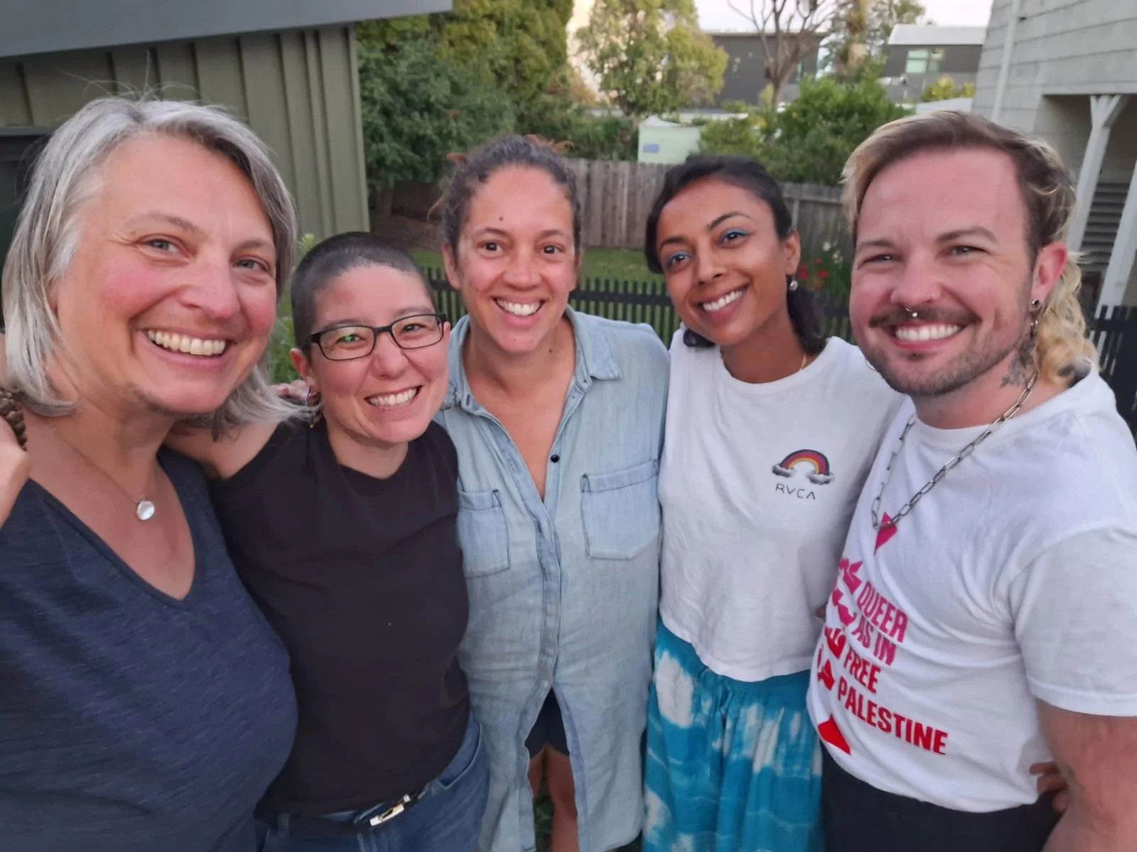 A group of five diverse friends smiling and standing close together outdoors in a backyard with trees and buildings in the background.