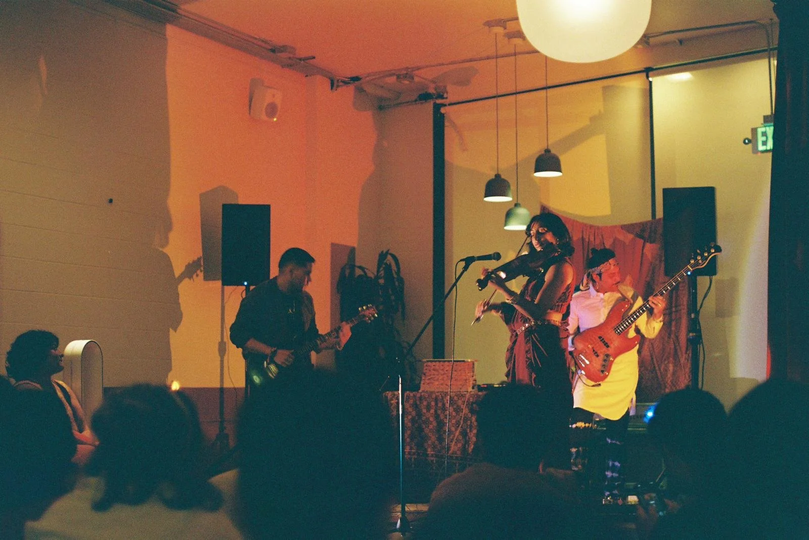 Musicians performing live on stage with colored lighting, including a woman playing the violin and singing, a man playing the bass guitar, and another man playing the electric guitar. Audience members are watching the performance.