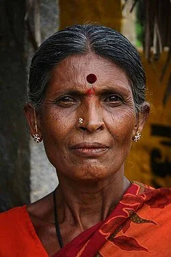 Close-up portrait of an elderly woman with a red bindi on her forehead, wearing jewelry and traditional attire, standing outdoors.