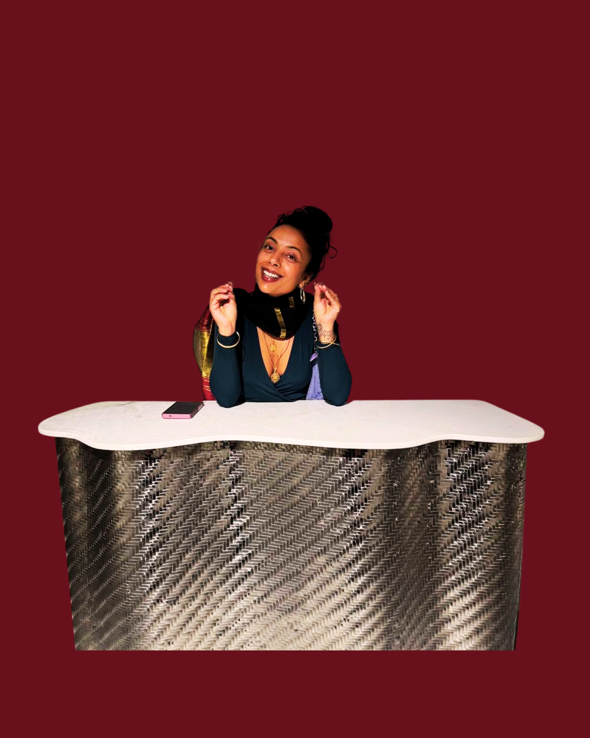A woman sitting at a white table with a wallet and phone, smiling and holding a scarf around her shoulders, against a dark red background.