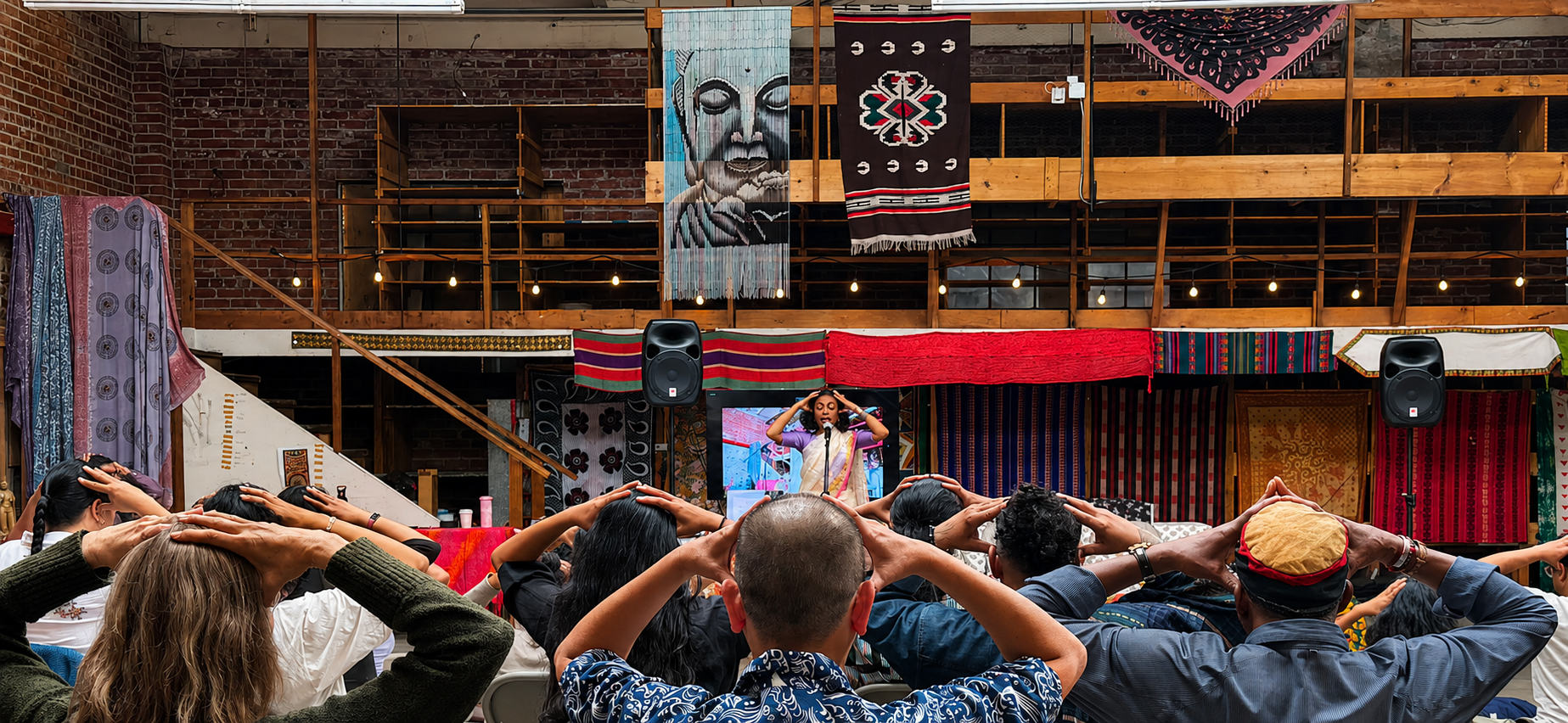 A group of people participating in a yoga or meditation class indoors, with a woman leading the session on stage. The room features exposed brick walls, colorful textiles and artwork hanging from the upper level, and stage lighting.