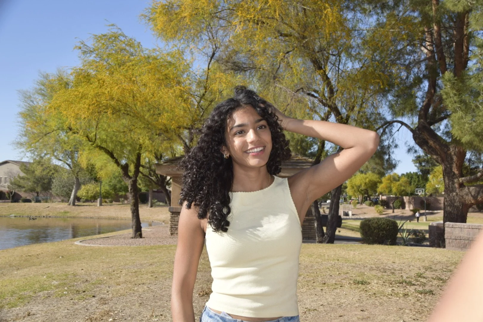A young woman with curly black hair smiling outdoors in a park on a sunny day, wearing a sleeveless yellow top and blue jeans, with trees and a pond in the background.