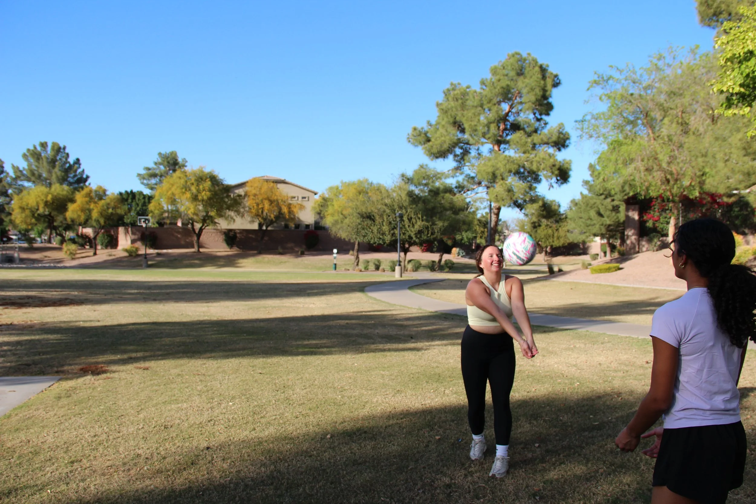 Two women playing volleyball outdoors in a park on a sunny day, with trees and a pathway in the background.