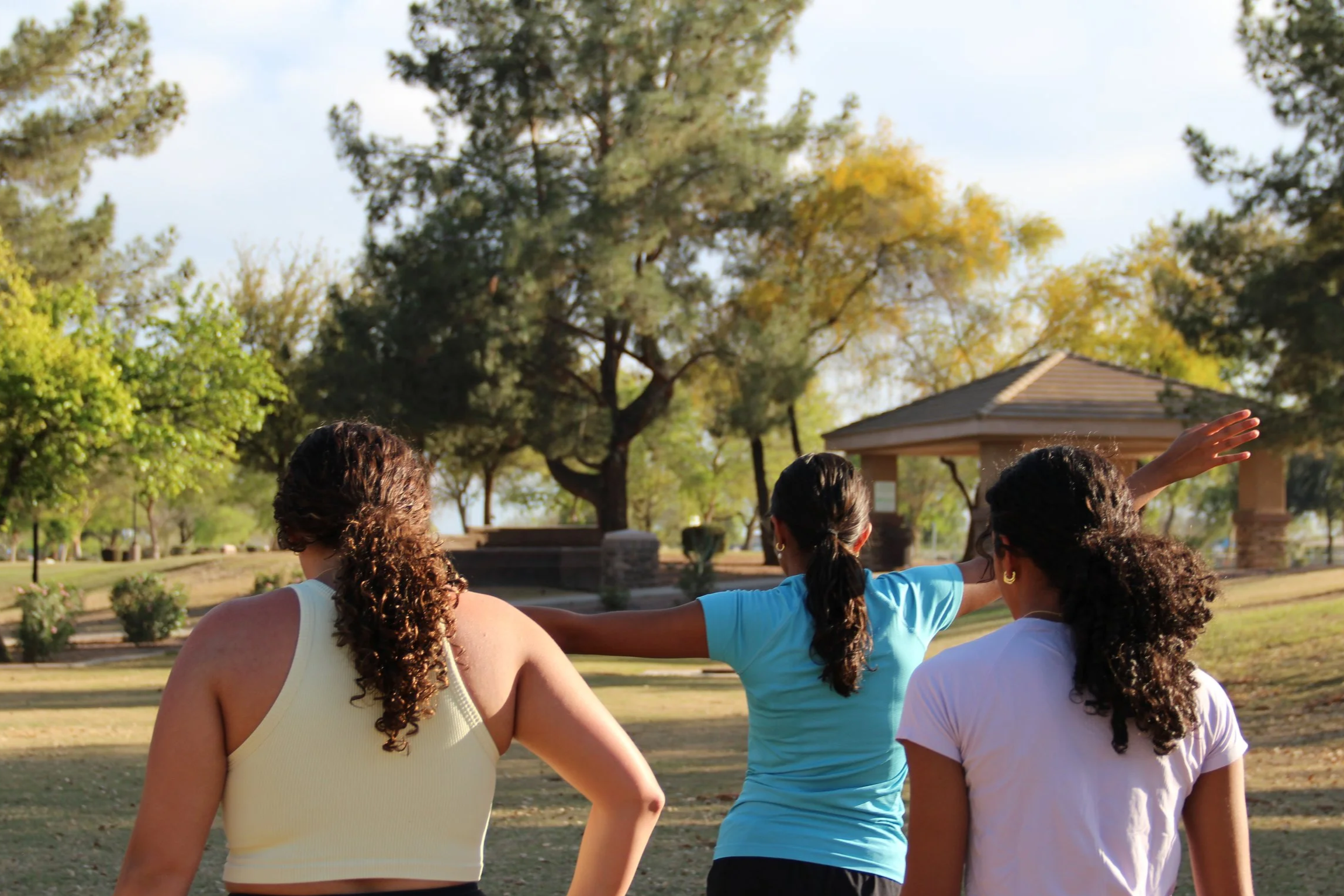 Three women standing outdoors in a park, with trees and a pavilion in the background, one woman is gesturing with her arm. Sunny weather.