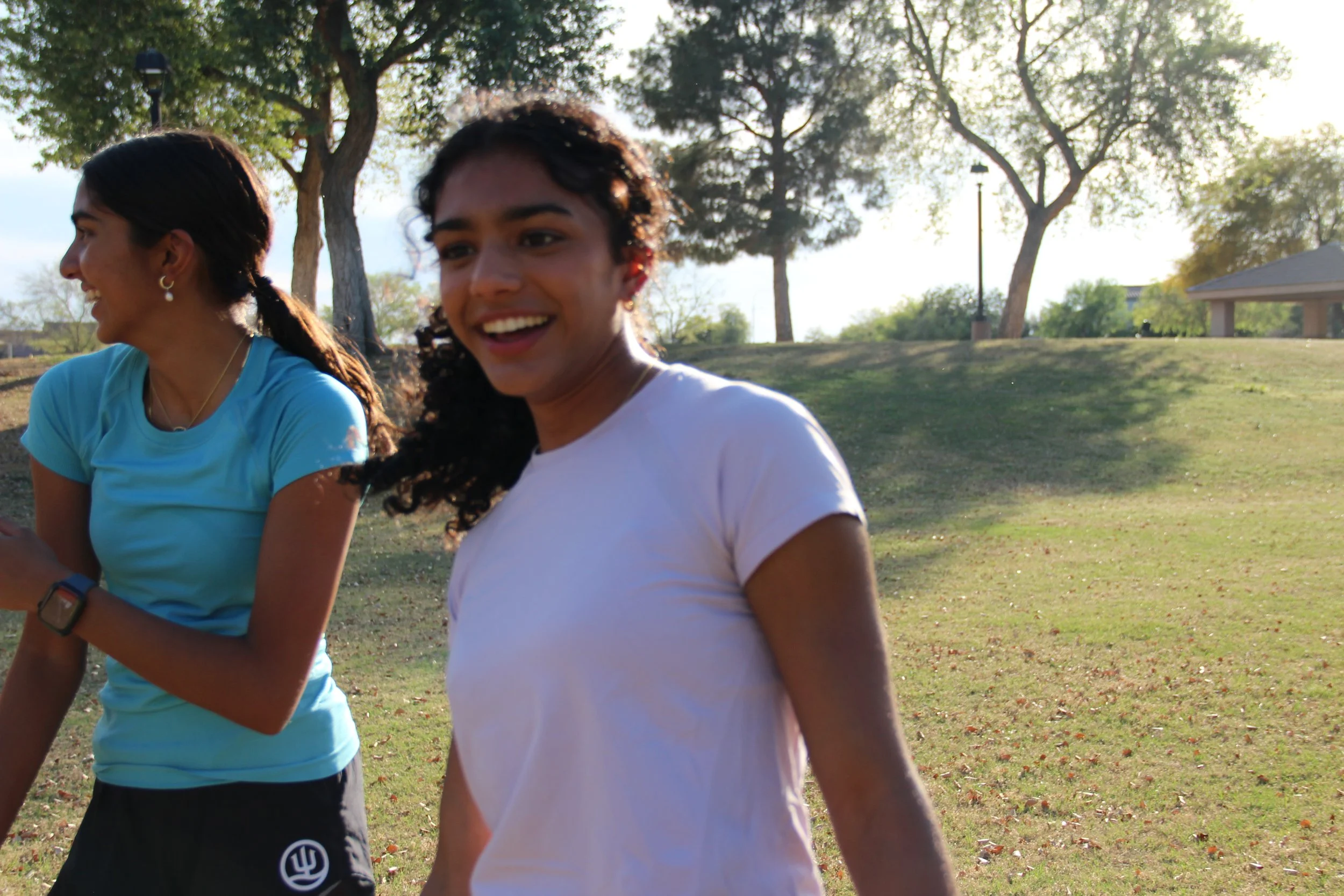 Two young women walking outdoors in a park during daytime, smiling and talking, with trees and a pavilion in the background.