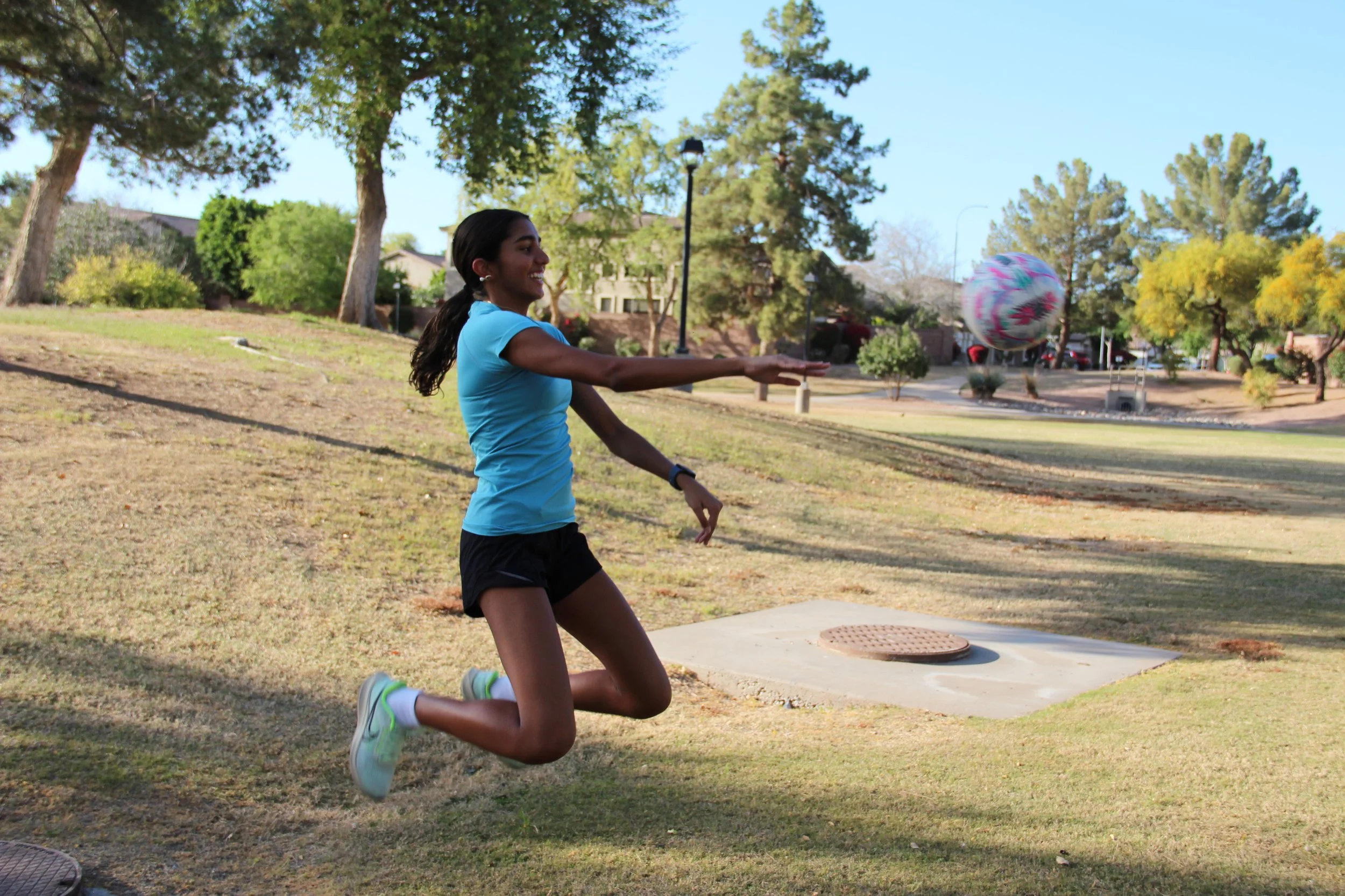 A young woman in a blue shirt and black shorts is jumping in a park to catch a colorful ball with a happy expression, surrounded by trees and open grassy areas.