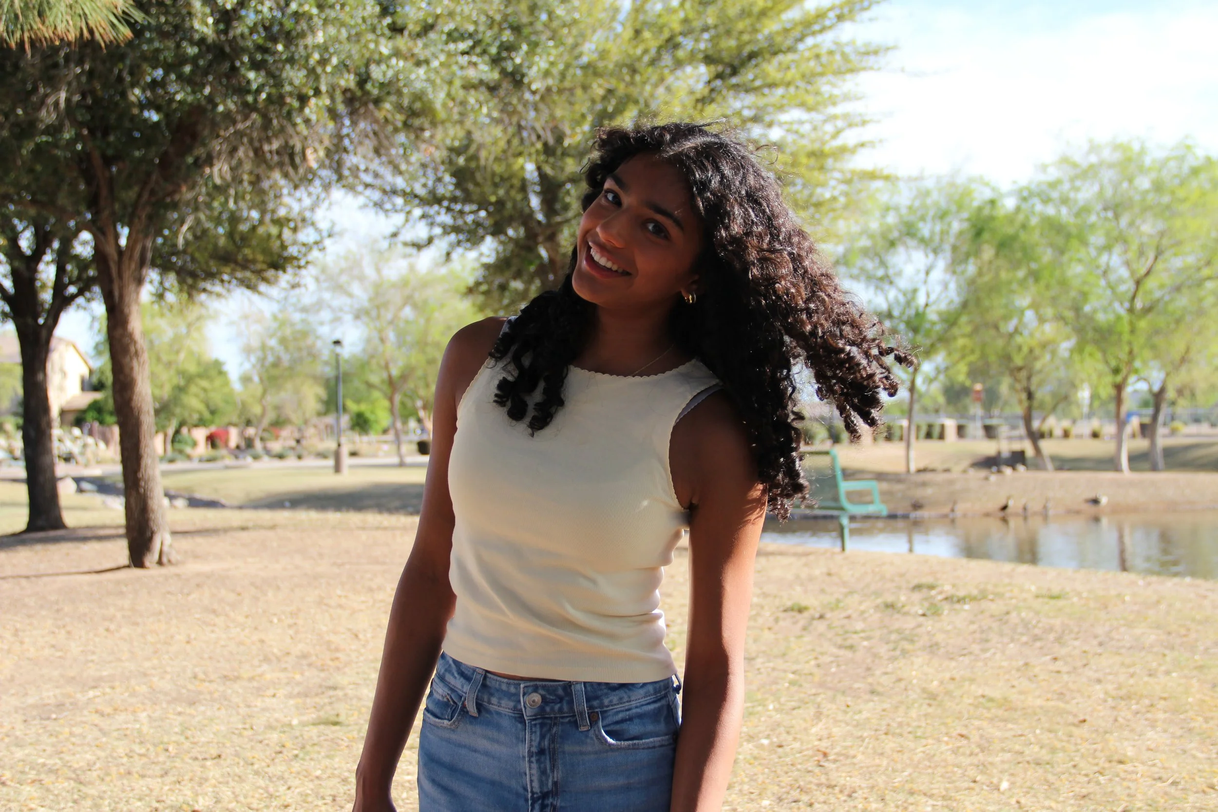 A woman with curly dark hair smiling and standing outdoors in a park with trees, a pond, and a bench in the background.