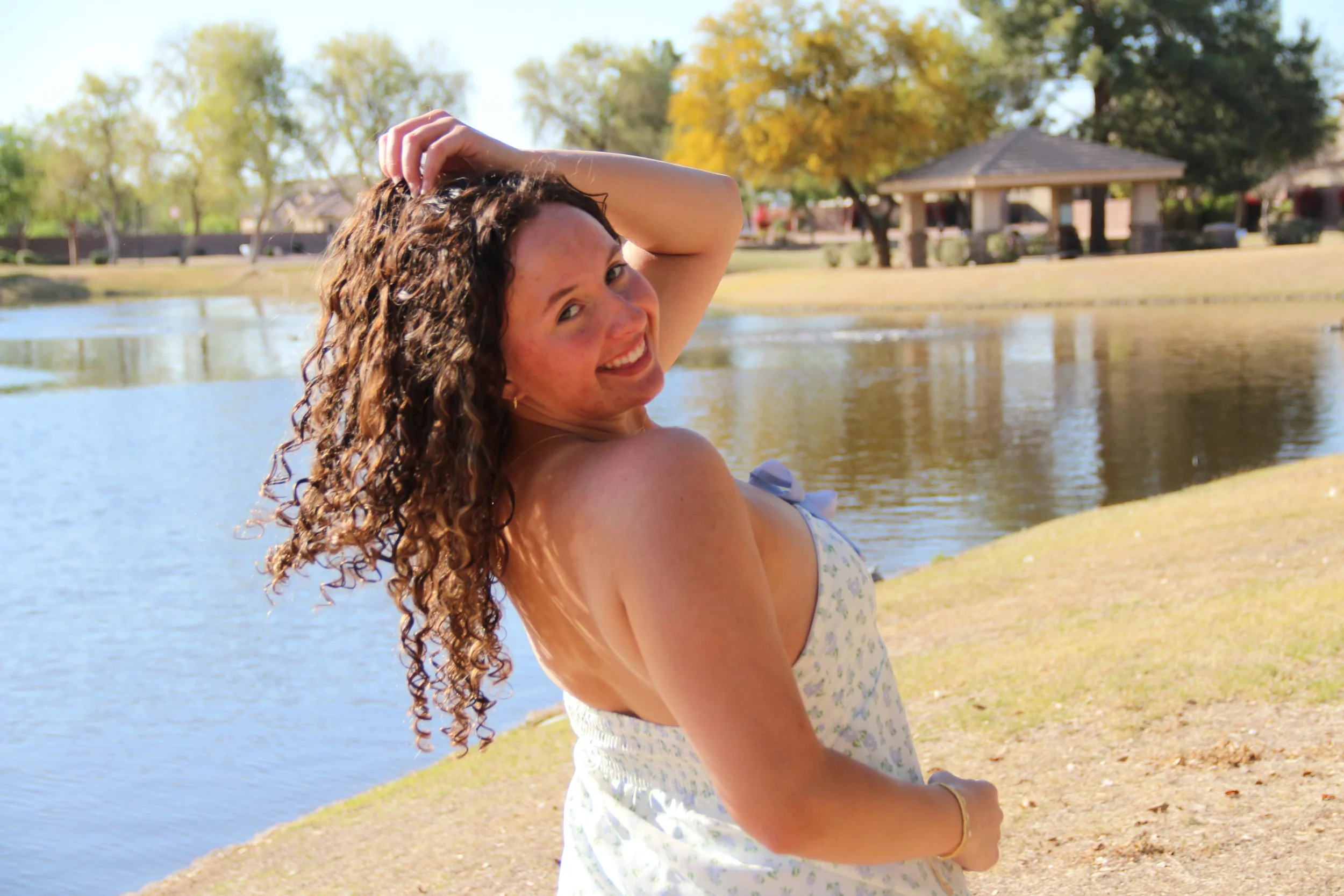 Smiling woman with curly hair, wearing a white floral dress, standing by a pond in a park on a sunny day.