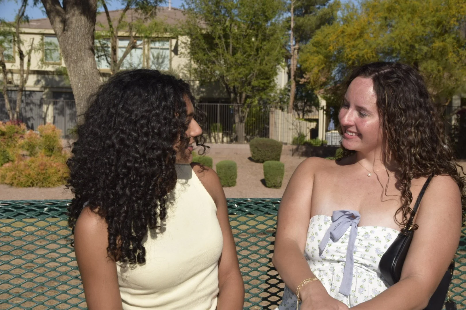 Two women sitting on a green park bench talking. One woman has dark curly hair, and the other has lighter curly hair. They are outdoors with trees and bushes in the background.