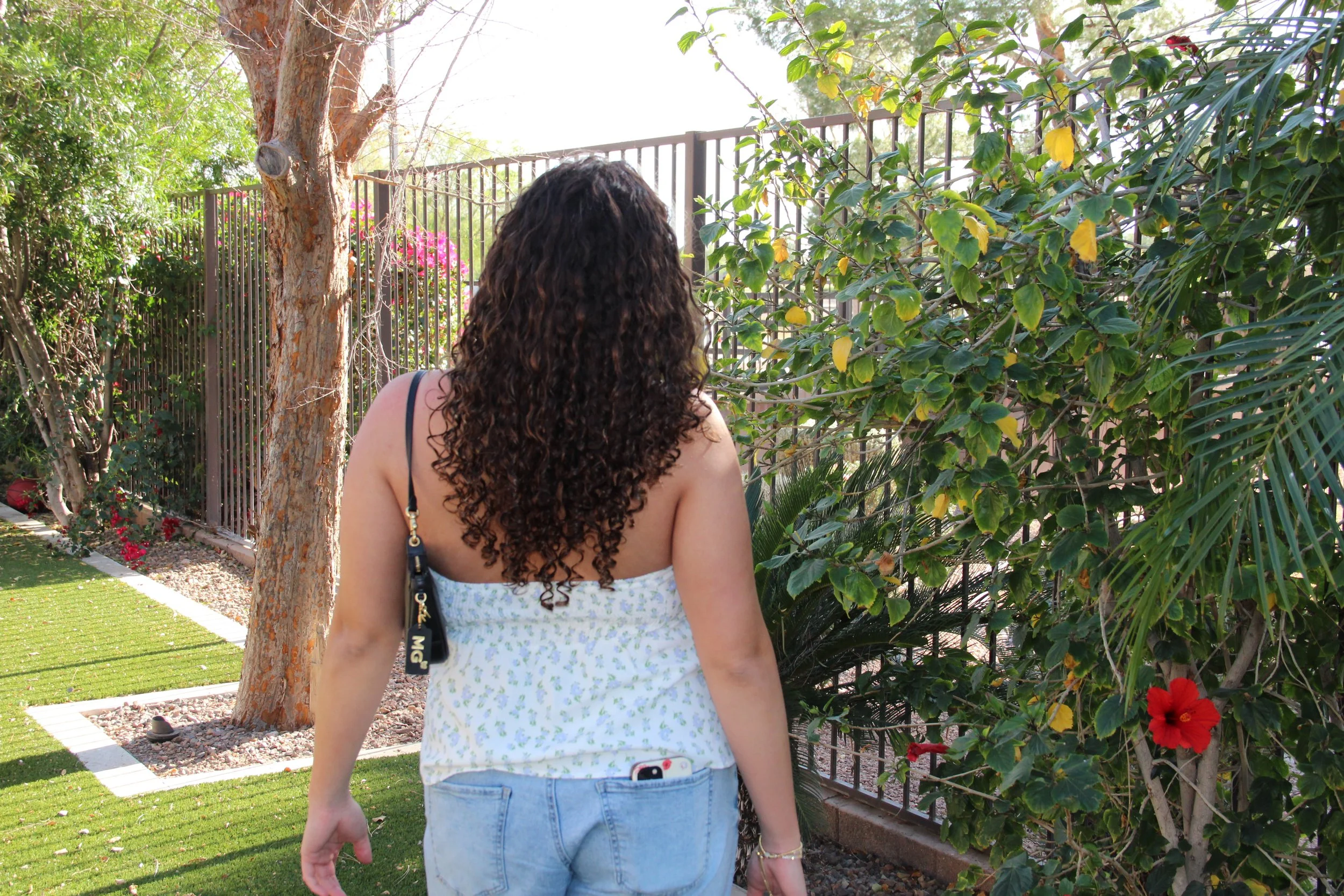 A woman with curly hair in a white tank top and jeans is standing outdoors near a tree and various green bushes with yellow and red flowers, facing away from the camera.