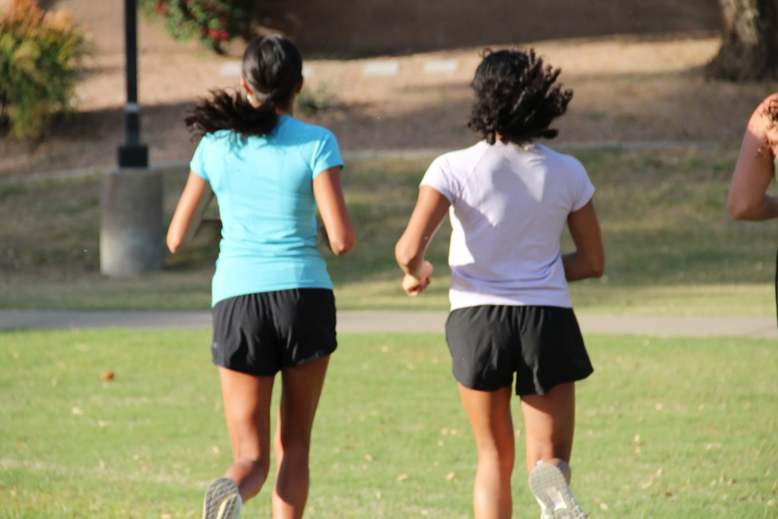 Two women jogging outdoors on a grassy park area, wearing athletic shorts and t-shirts, with trees and a pathway in the background.