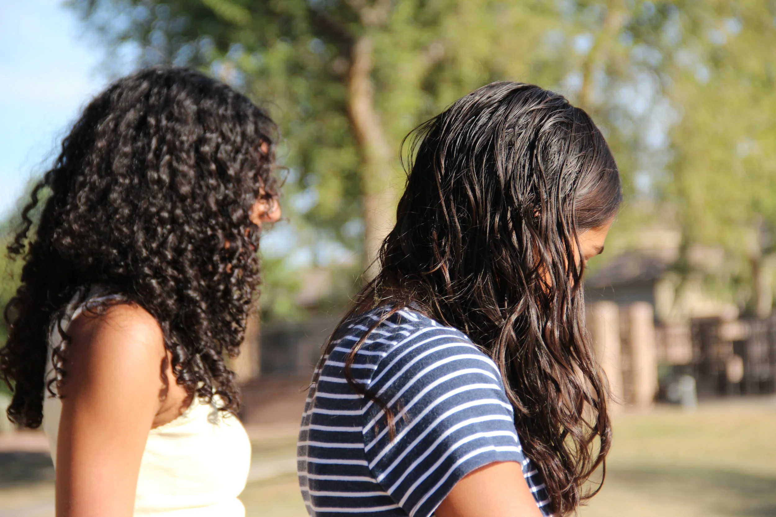 Two young women with long dark hair, one with curly hair and the other with wavy hair, standing outdoors with trees and a fence in the background.
