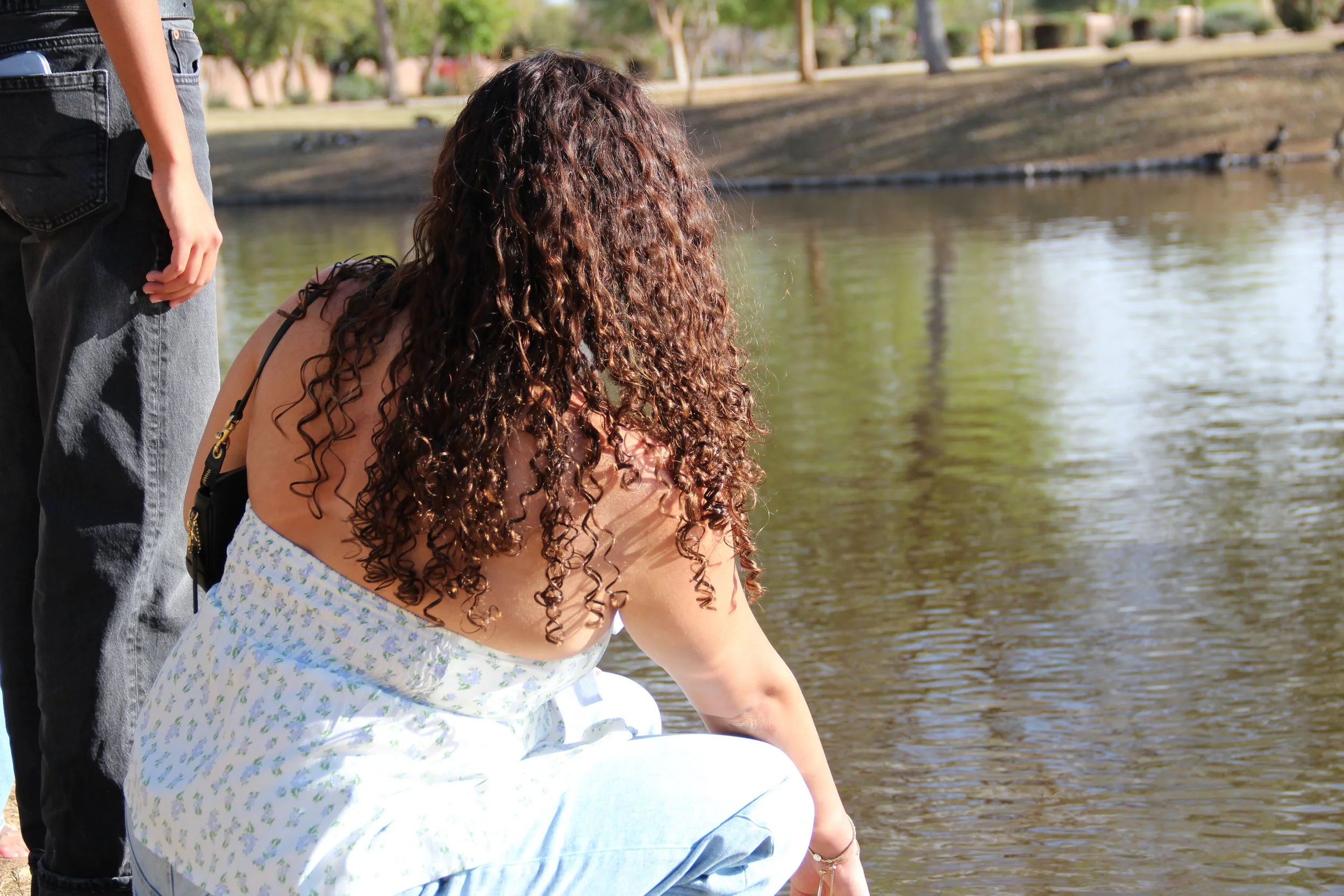 A woman with long, curly hair in a white dress squatting by a body of water, with a person standing nearby.