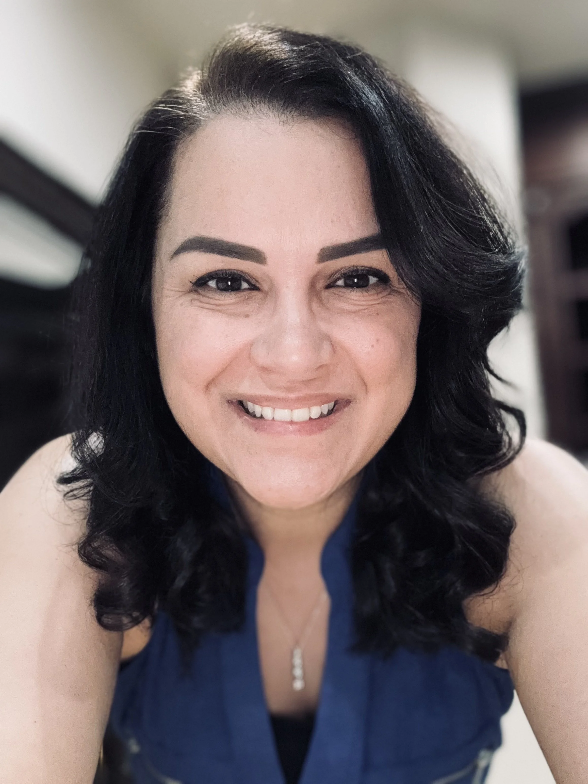 Close-up of a woman with black hair and smiling face, wearing a blue top and silver necklace.