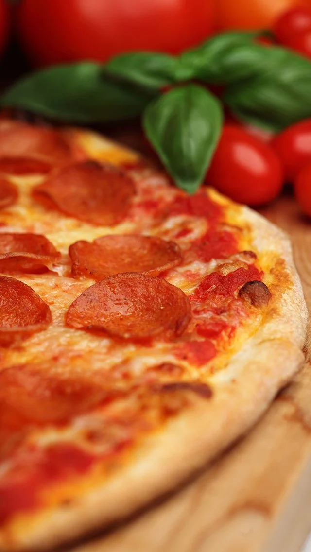 Close-up of a pepperoni pizza with fresh basil leaves and cherry tomatoes in the background.