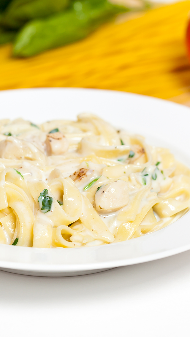 A bowl of creamy chicken Alfredo pasta with green herbs, served on a white plate. In the background, a colorful display of vegetables.