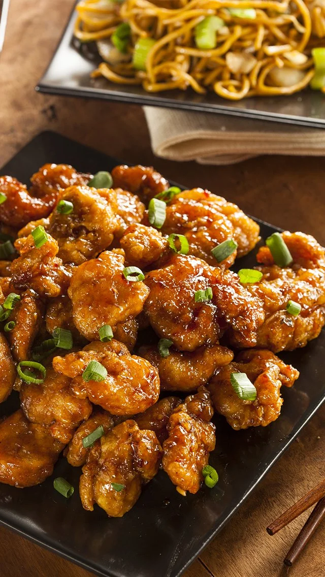 Sweet and sour chicken with chopped green onions on black rectangular plate, with a side of stir-fried noodles and vegetables in the background.